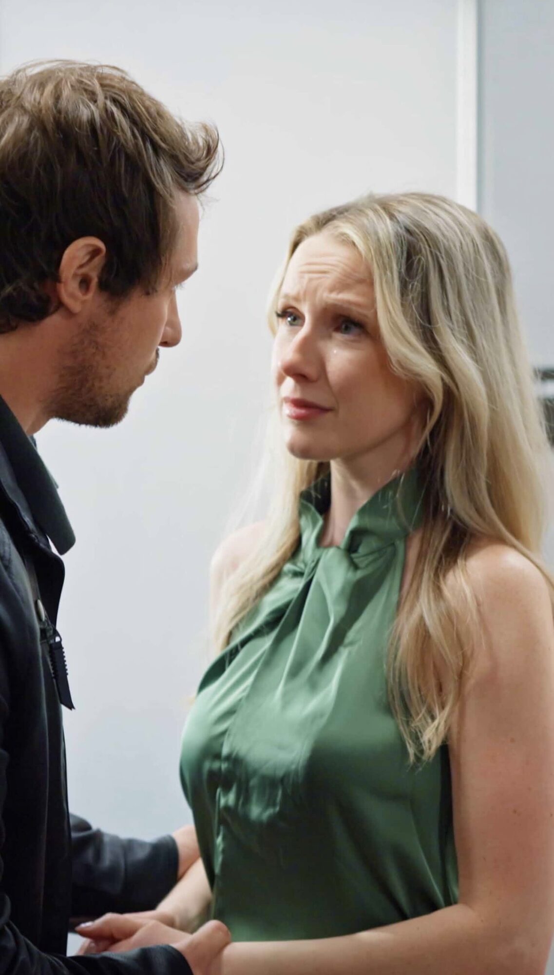 A man and woman face each other indoors, engaging in a serious conversation, with neutral background and natural lighting.