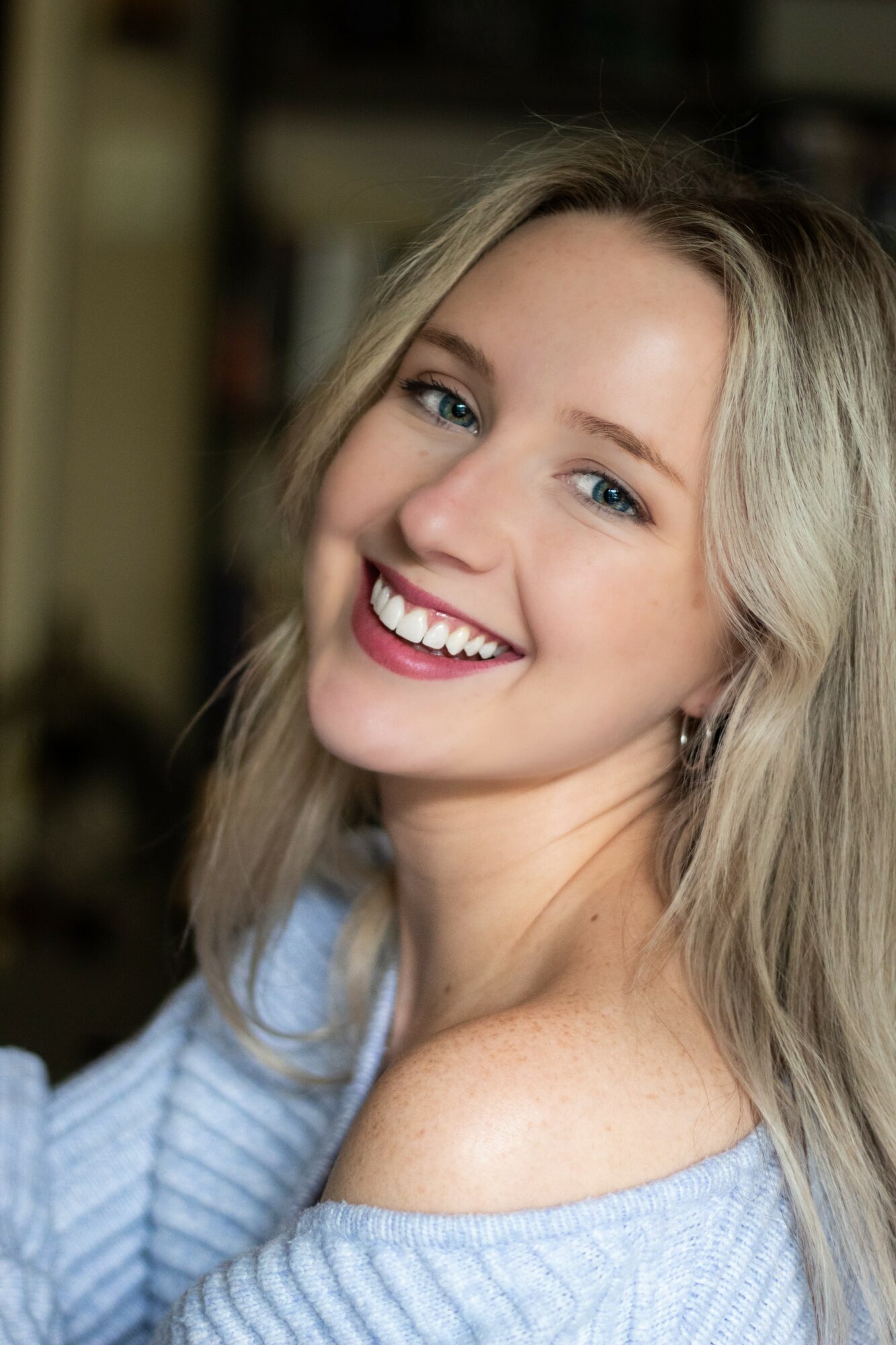 Young woman with blonde hair smiling, wearing a light blue off-shoulder sweater, indoors with blurred background.