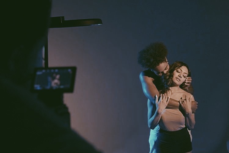 Two women embrace on a dark stage, one with curly hair and the other with straight hair, with a person filming them in the foreground.