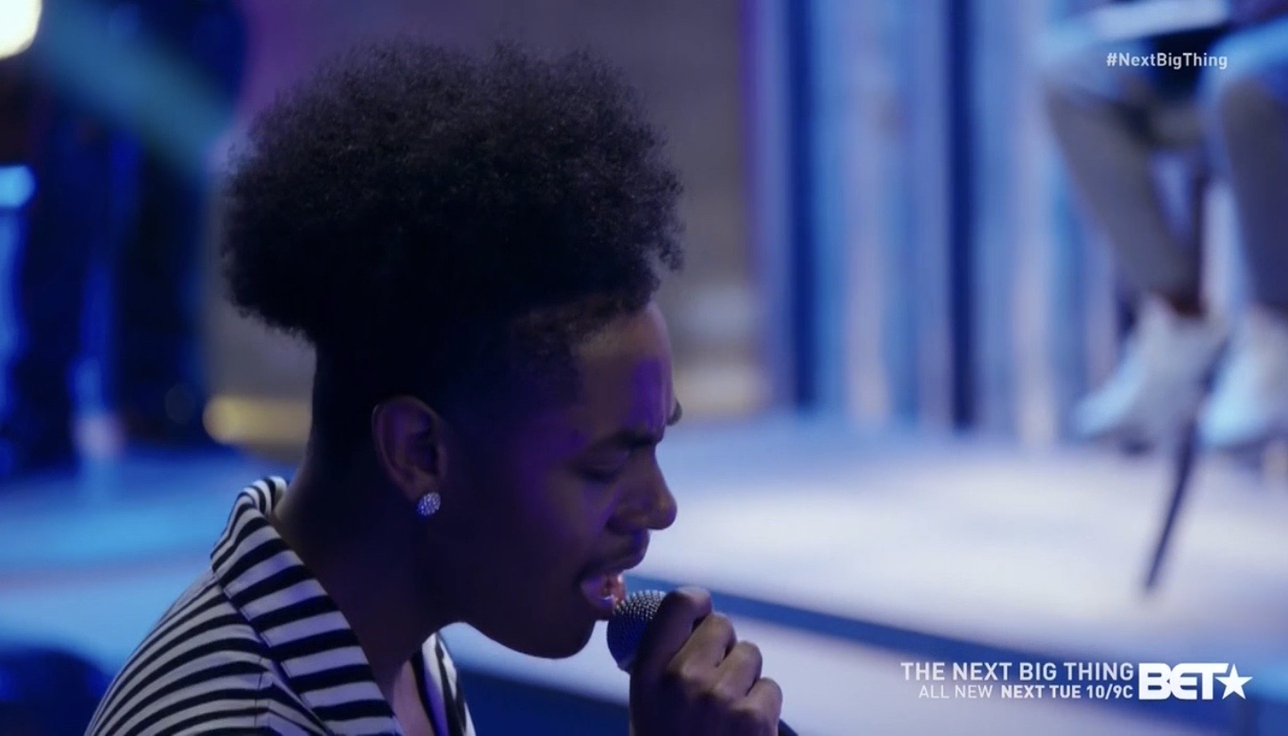 Woman with curly hair singing into a microphone, wearing a striped shirt, in a blue-lit setting.