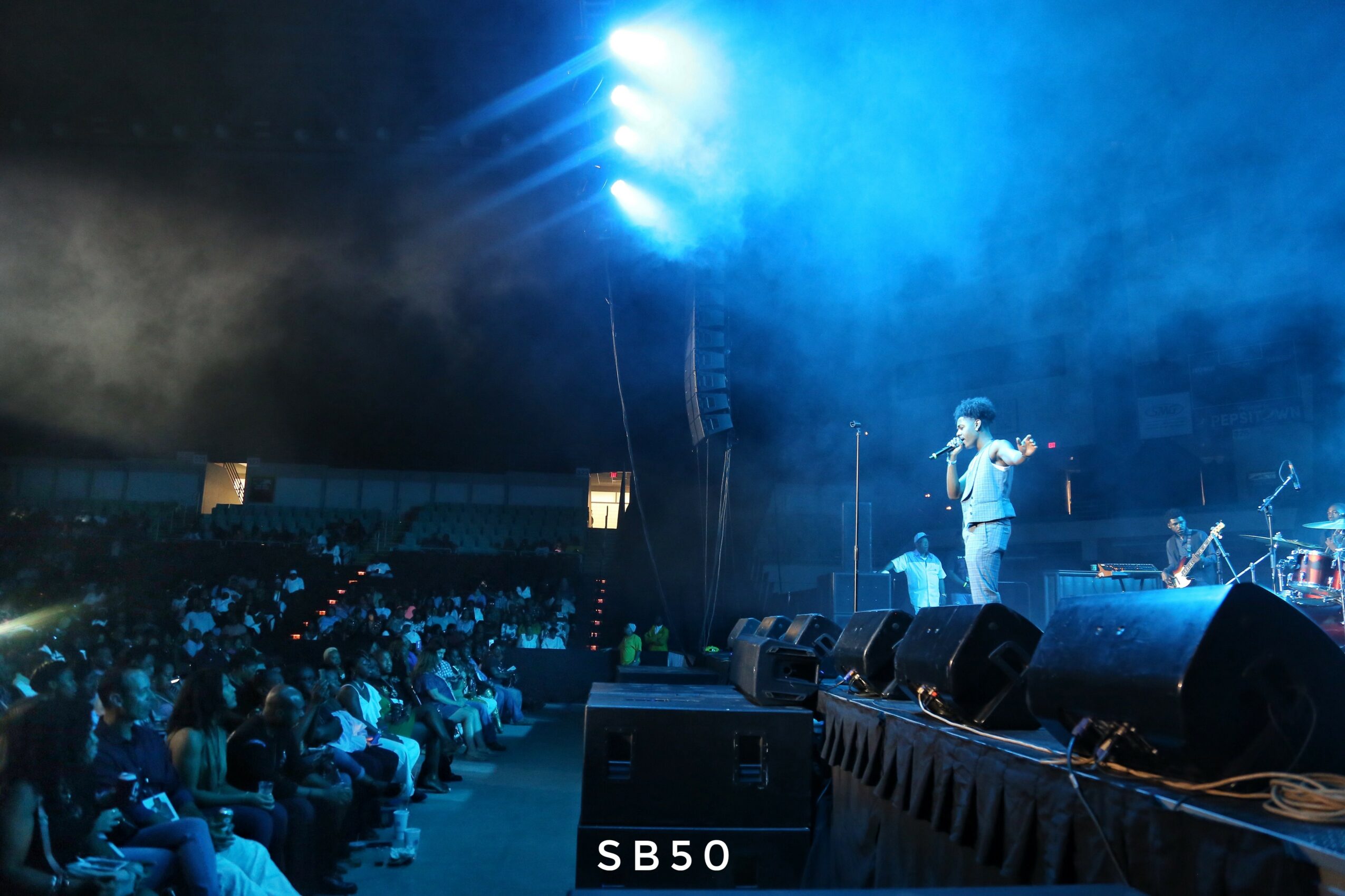 Performer on stage with audience in a large indoor venue, blue lighting, smoke effects, and sound equipment visible.