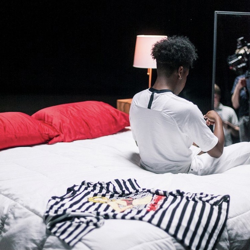 Person with curly hair sitting on bed, facing mirror, with red pillows and striped clothing in a dimly lit room