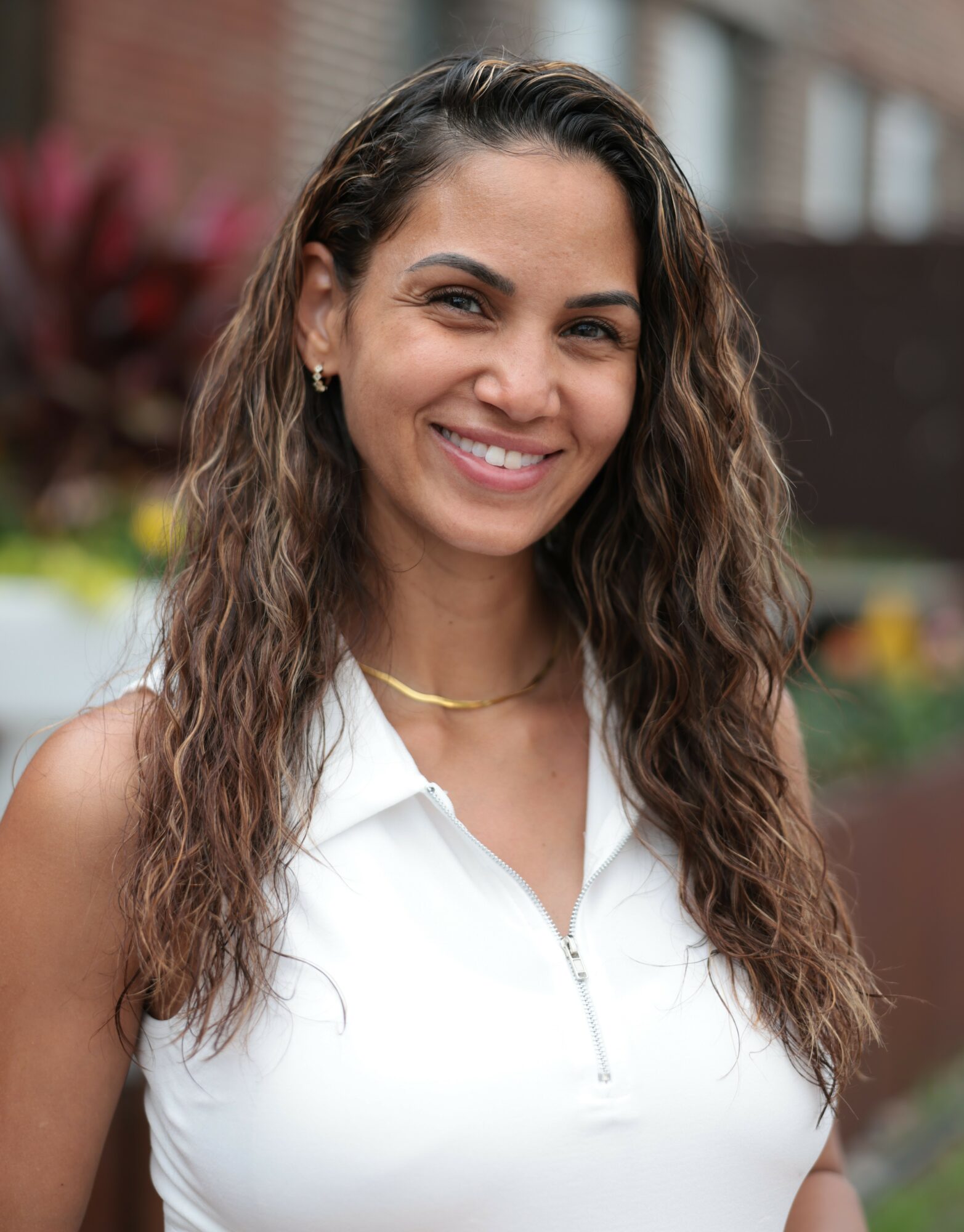 Smiling woman with long wavy hair wearing a white sleeveless top outdoors, blurred background of plants and building.