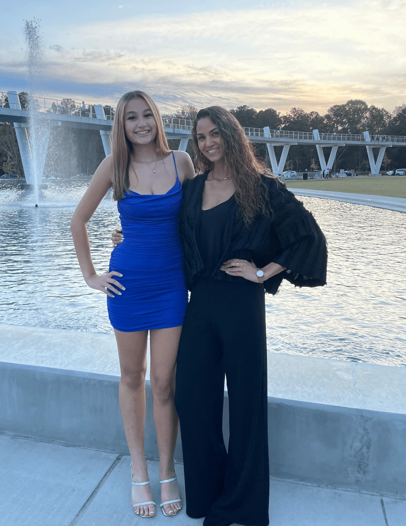 Two women standing by a fountain with water jets, smiling, during sunset, one in a blue dress, the other in black pants and top.