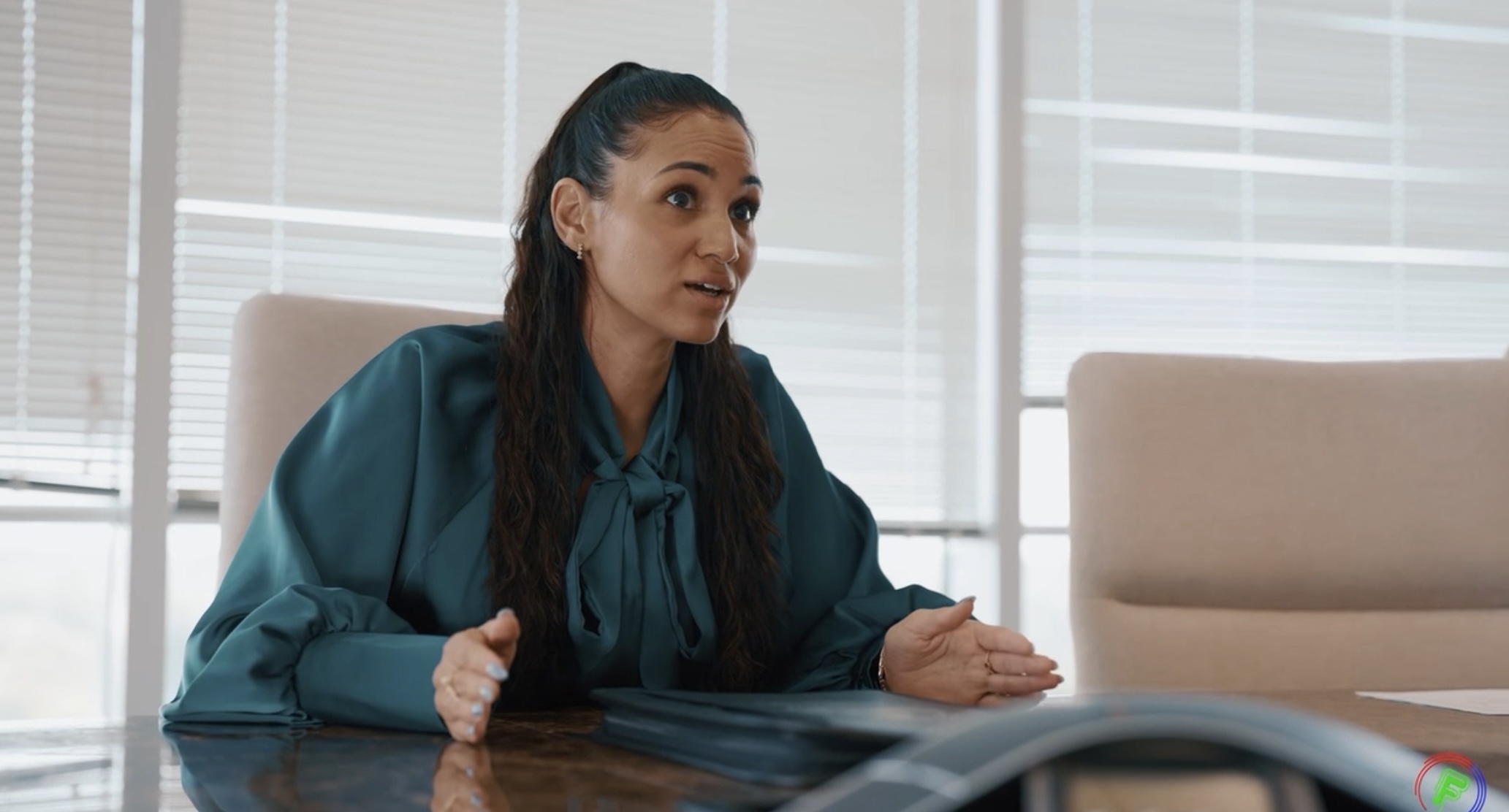Woman with long dark hair in a teal blouse sitting at a table in a bright room with large windows.