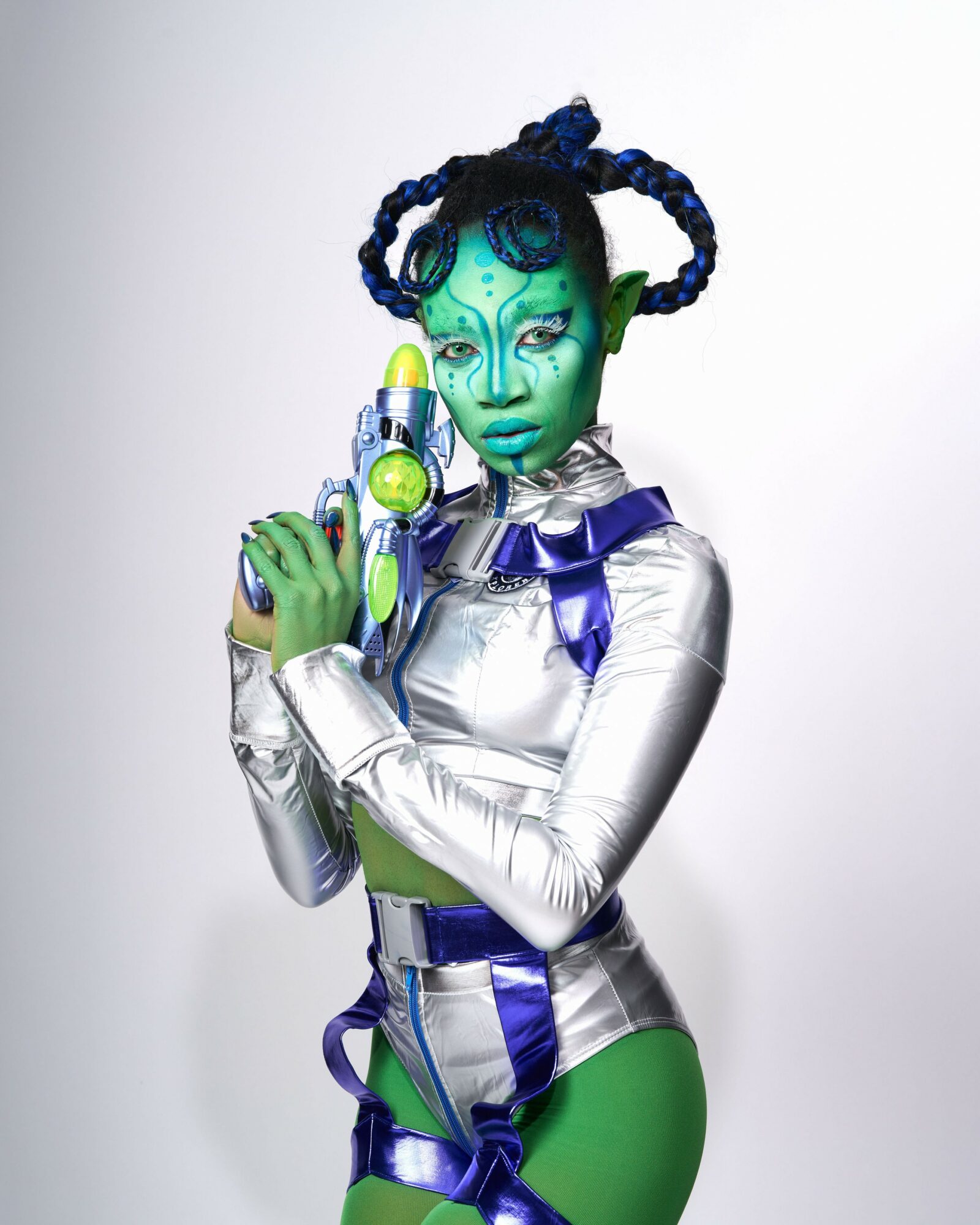 Person with green skin, elaborate blue hair, metallic silver outfit, holding a futuristic device, against a plain background.