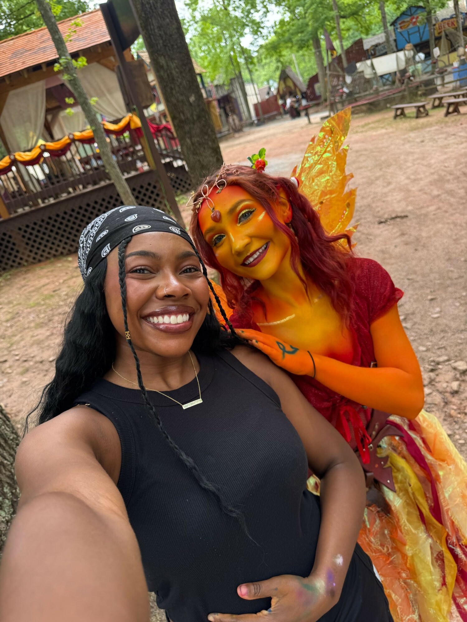 Two women smiling, one in colorful costume with red hair, the other in black top with glasses, outdoors.