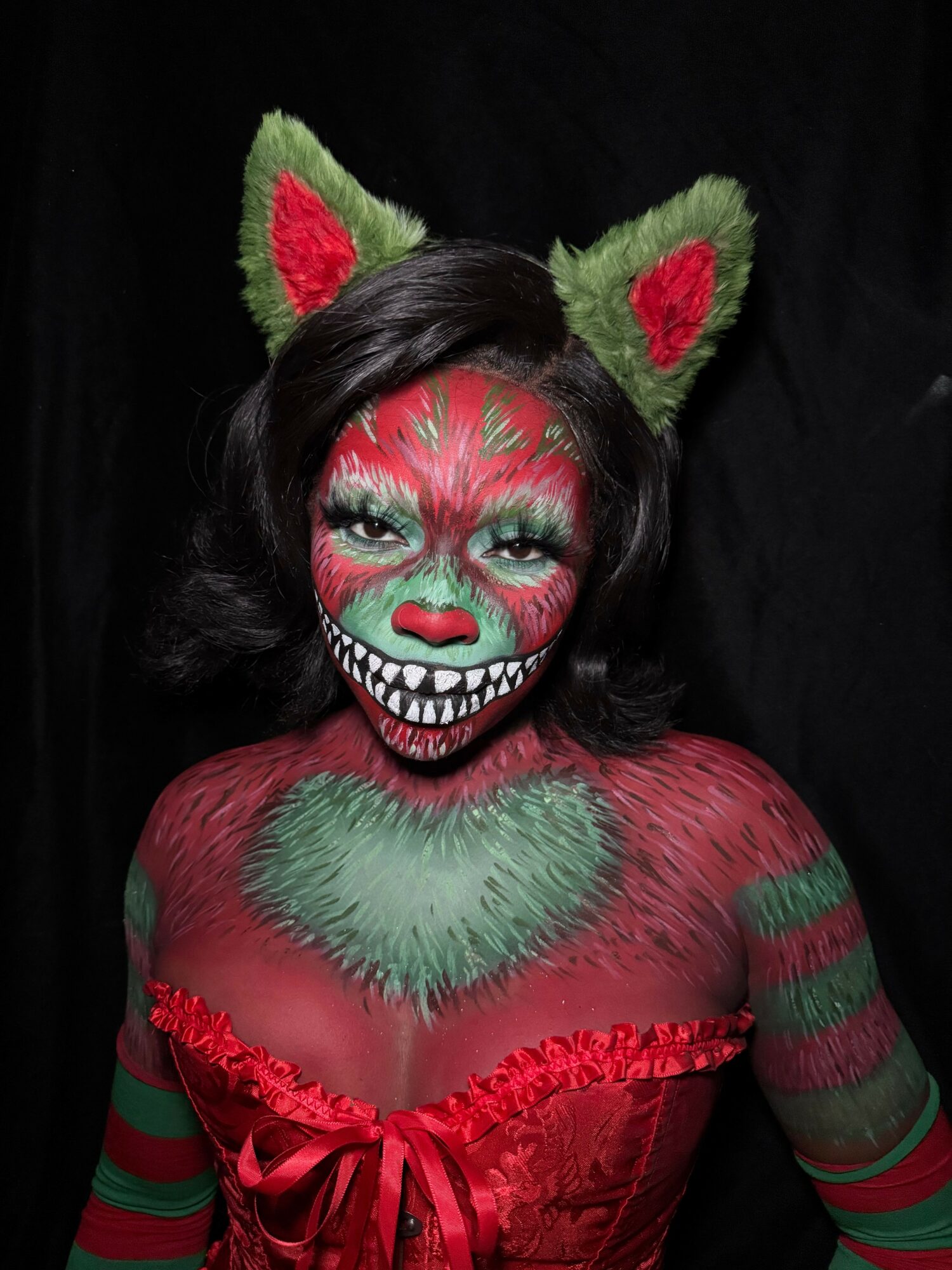 Person with green and red face paint, cat ears headband, and colorful costume, lying on black background.