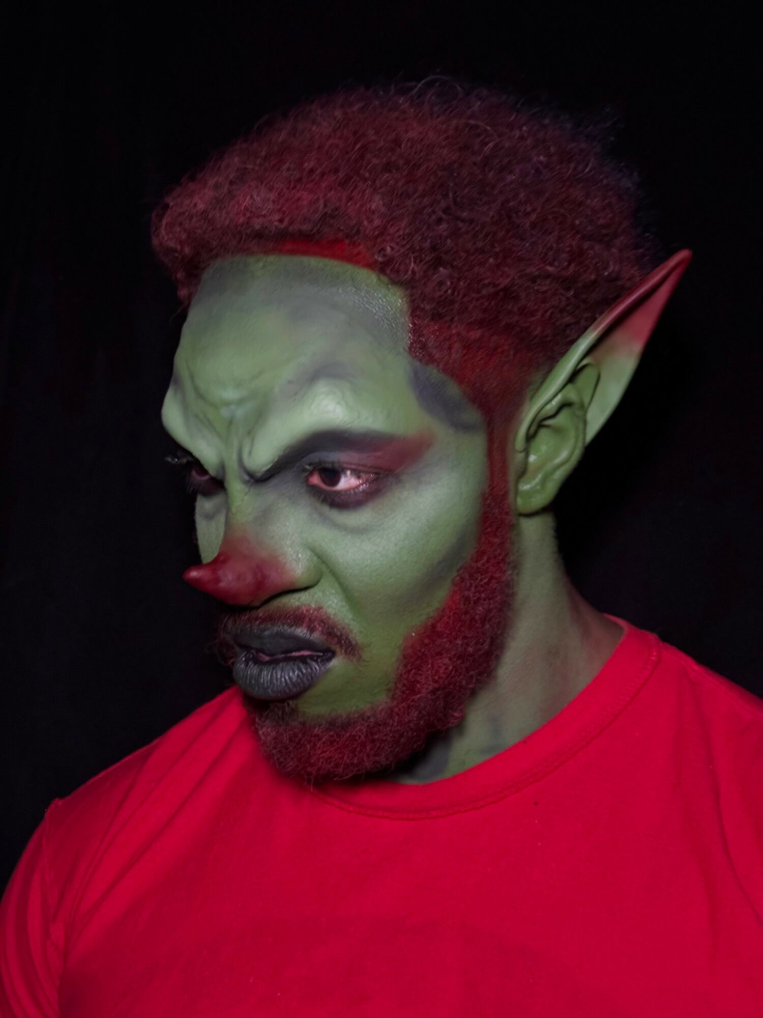 Person with green face makeup, pointed elf ear, red hair, and red shirt, looking to the side with a serious expression.