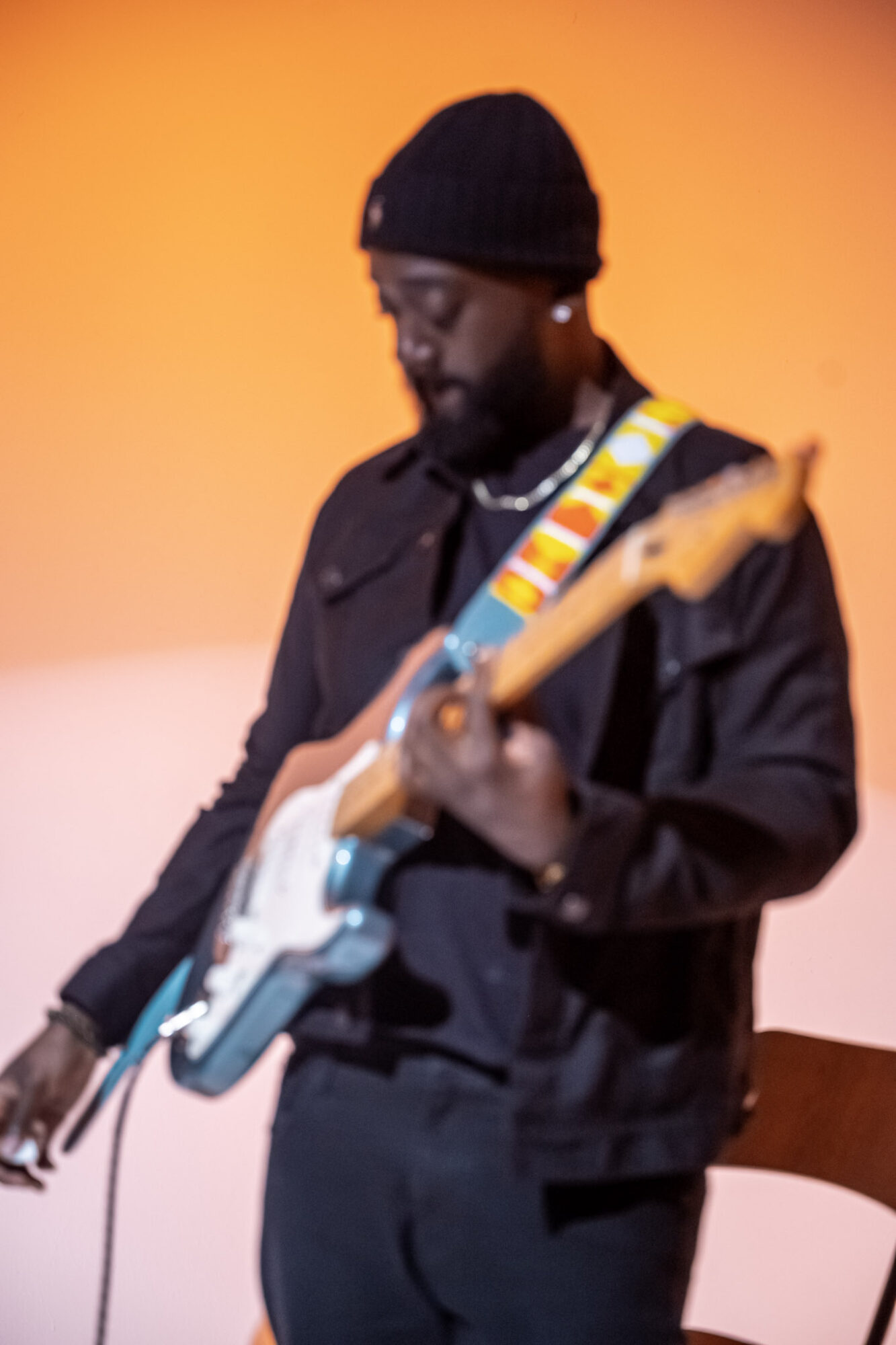 Person playing electric guitar, wearing a black beanie and jacket, against an orange background.