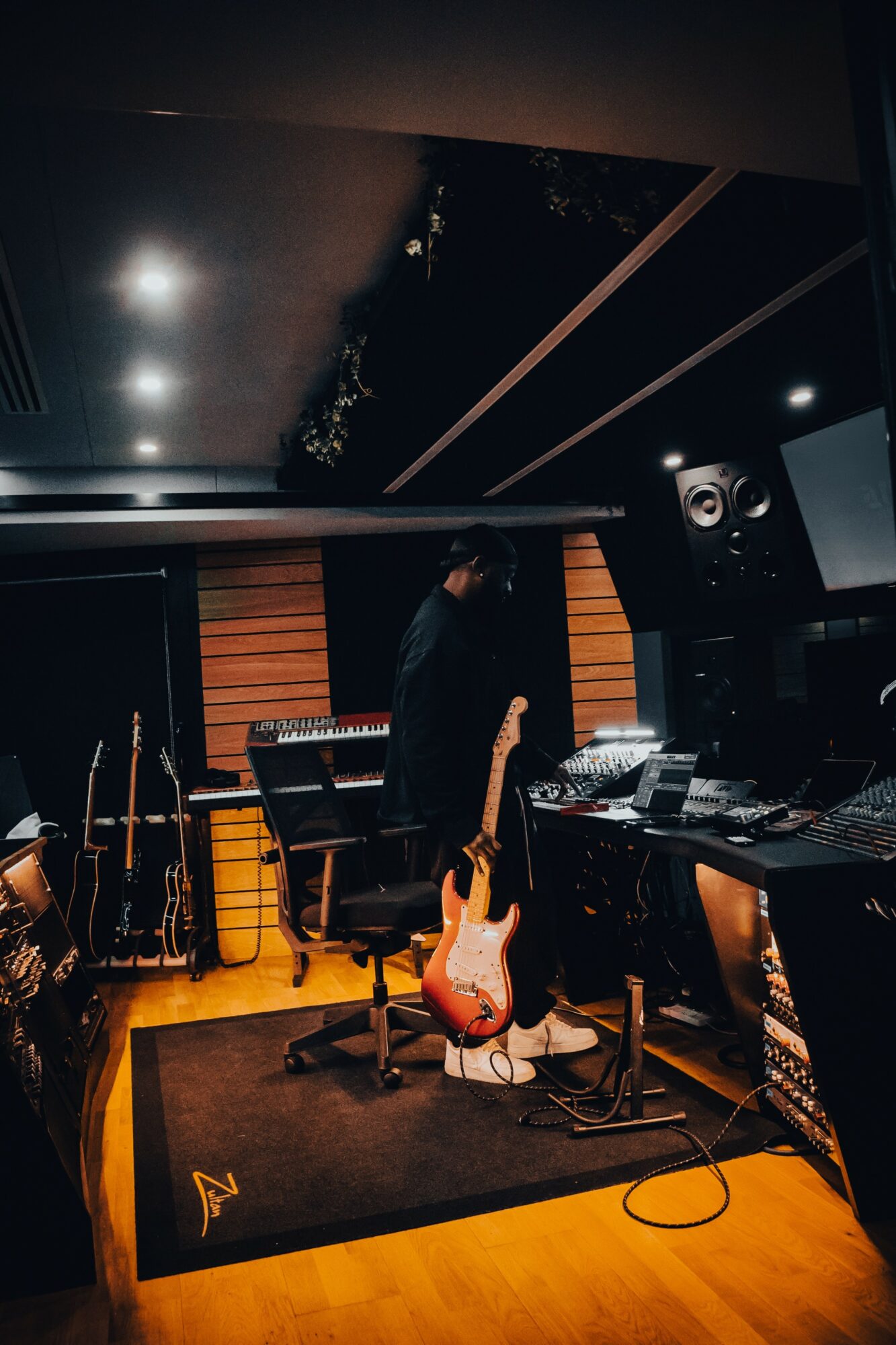 Person standing in a music studio with guitars, keyboards, and audio equipment, facing away from the camera, dimly lit room.