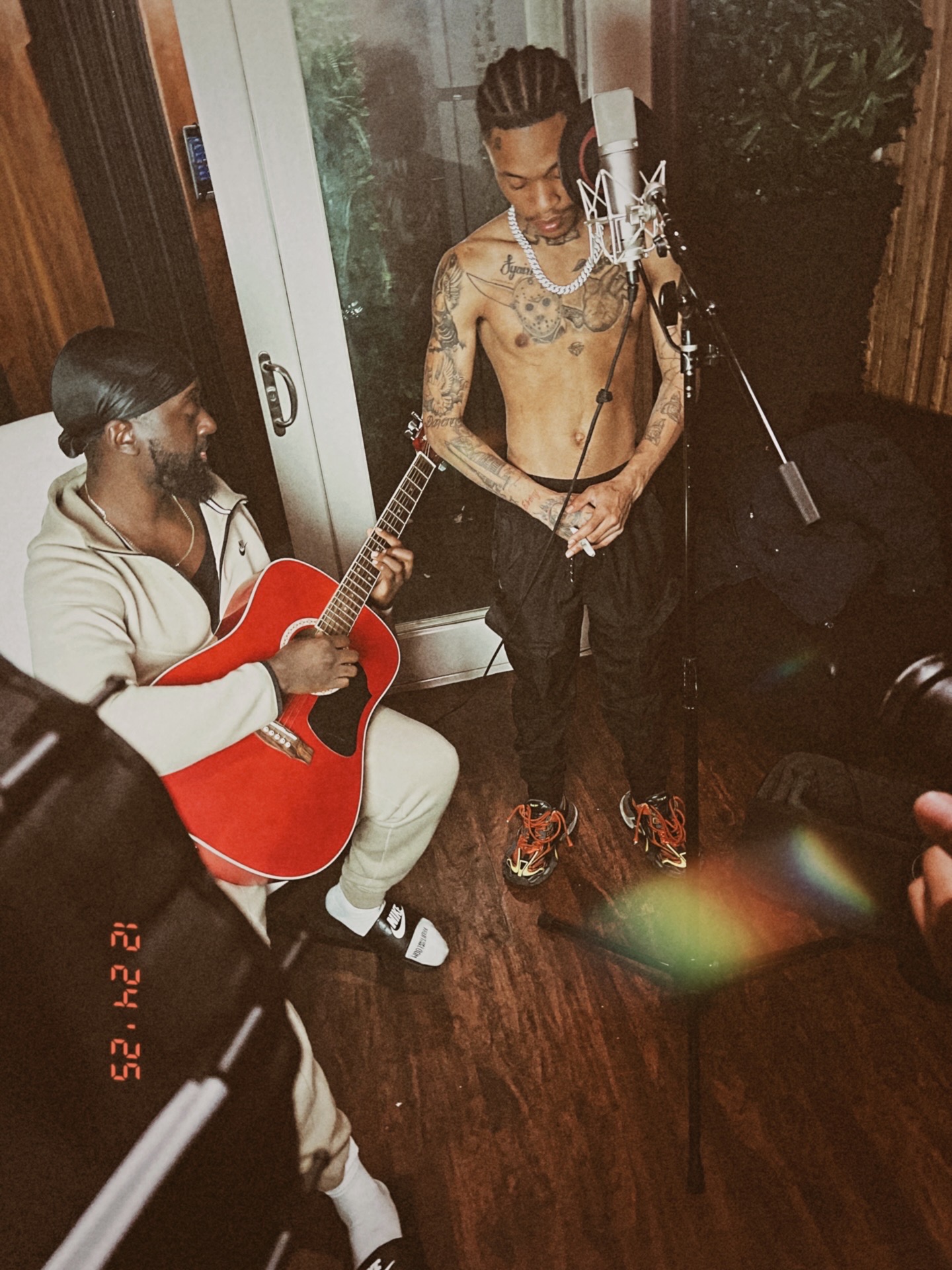 Two men in a recording studio, one shirtless with tattoos and the other playing a red guitar, standing near a microphone.