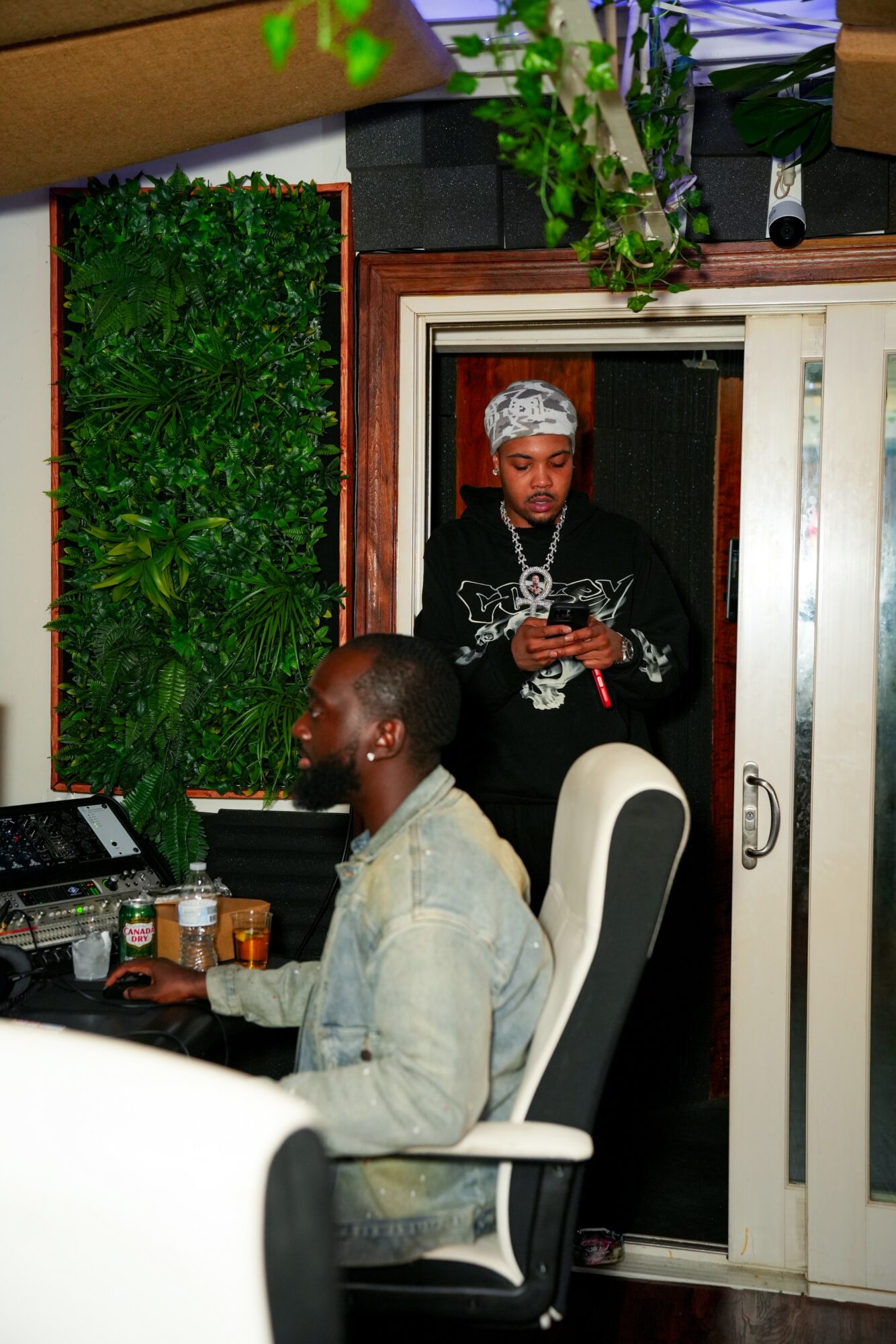 Two men in a room, one sitting at a desk and the other standing near a doorway, with green plants on the wall.