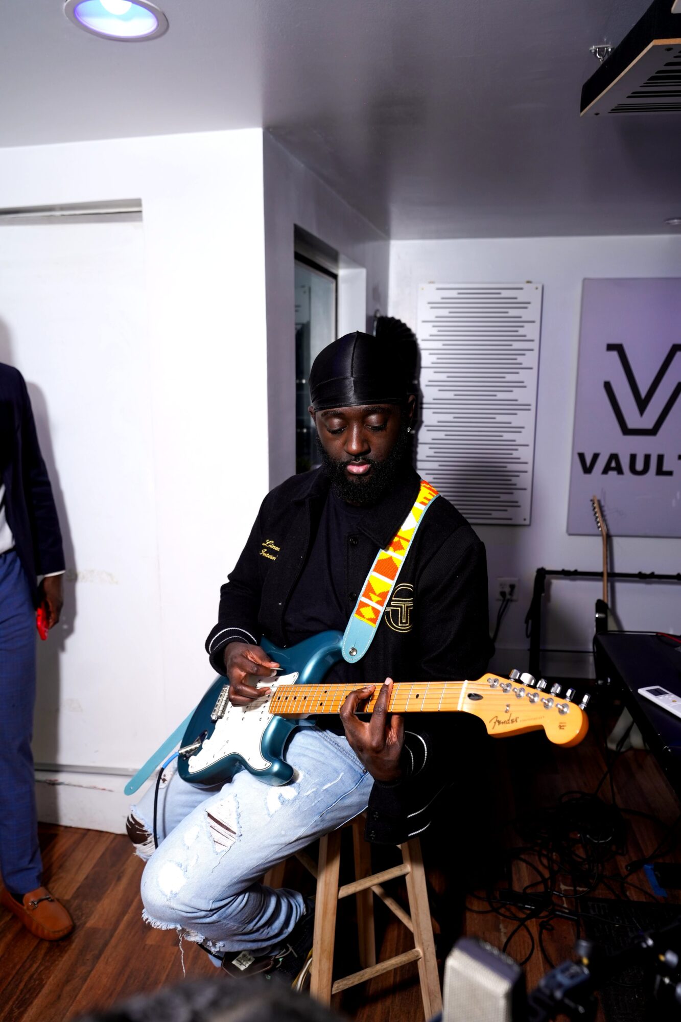 Person with dark hair and glasses playing an electric guitar in a room with white walls and a VAULT sign.