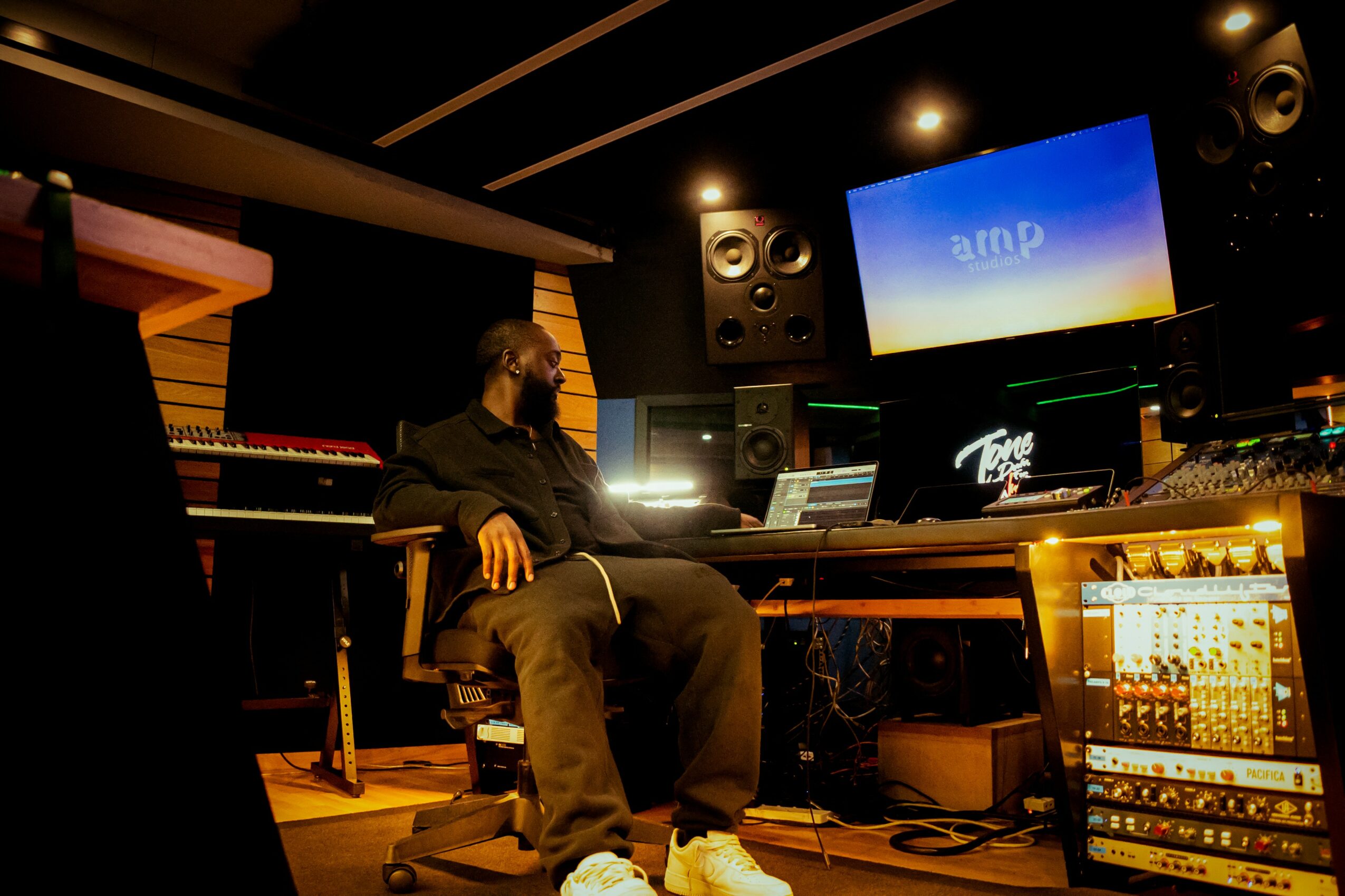 Person sitting in a music studio with equipment, monitors, and a large screen displaying blue background, in a dimly lit room.