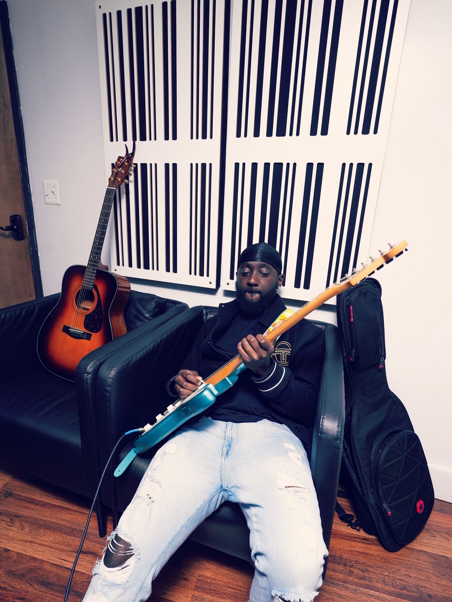 Person sitting on a black couch holding a blue electric guitar, with an acoustic guitar and guitar case behind them.