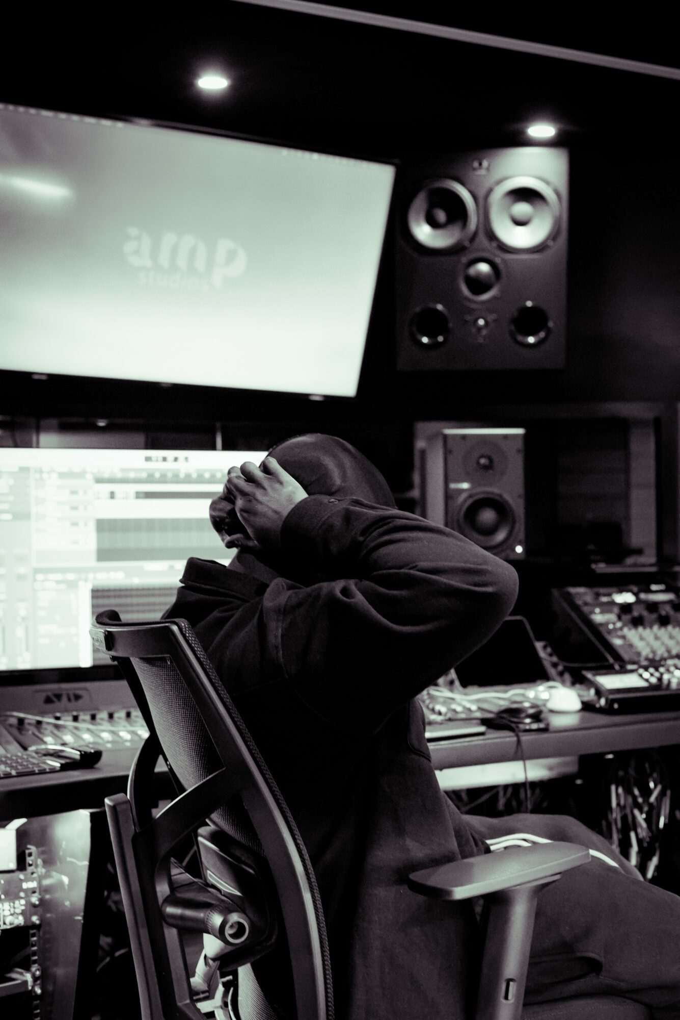 Person sitting in a music studio with headphones, surrounded by audio equipment and speakers, facing a large screen.