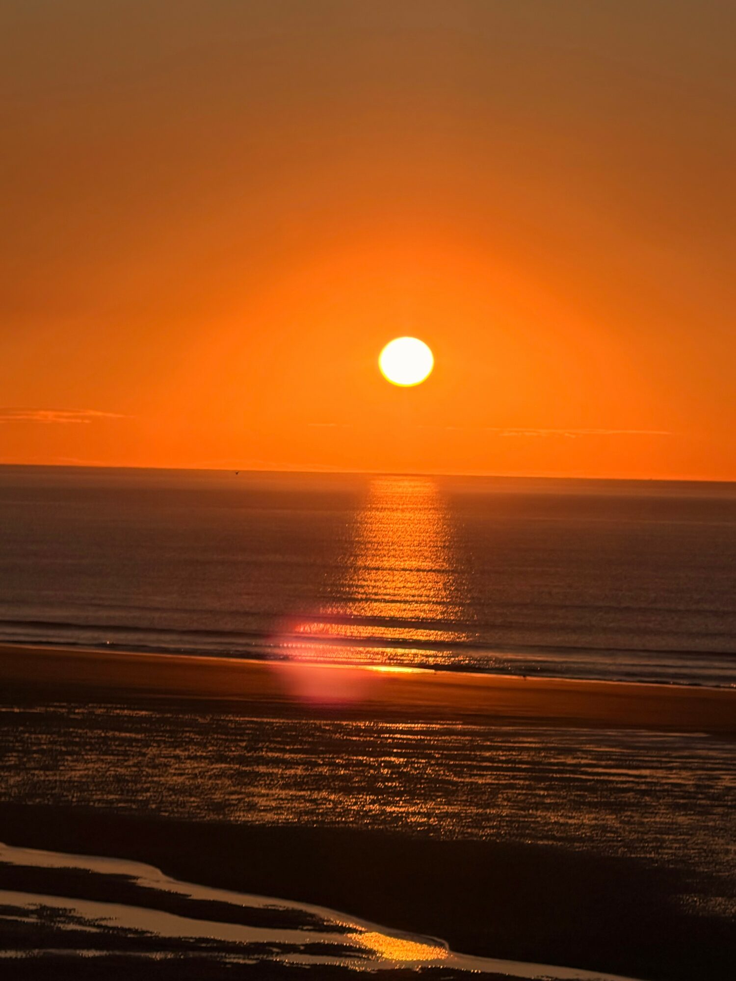 Sunset over the ocean with orange sky, reflecting on water, and a sandy beach in foreground.