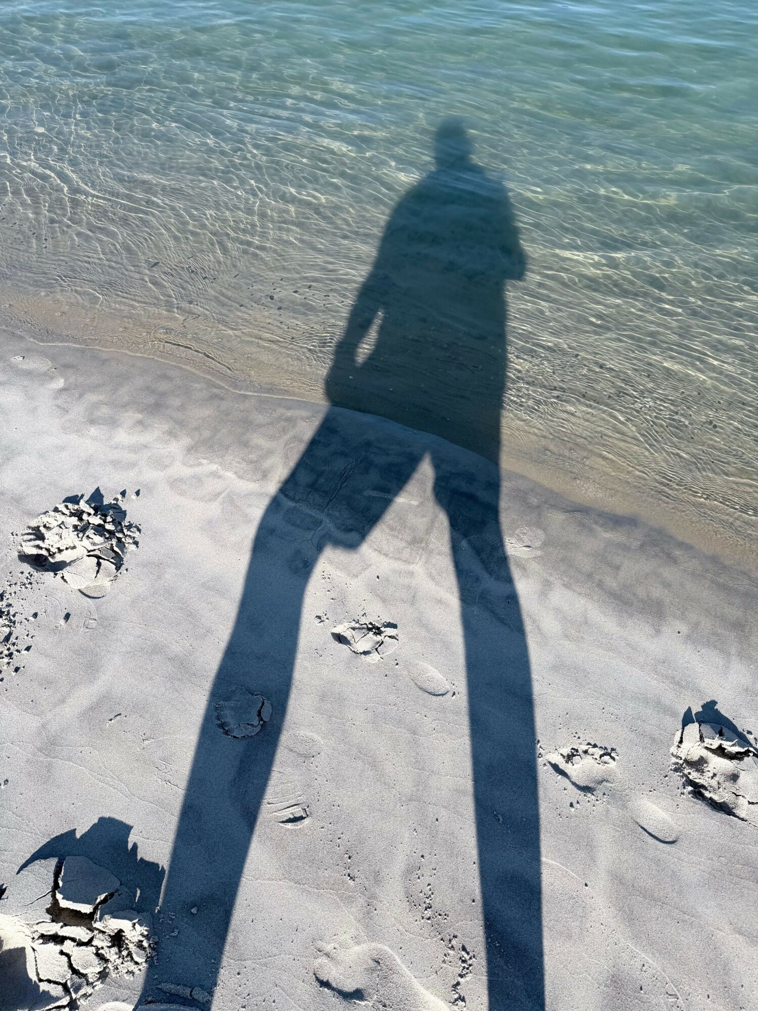 Shadow of a person on a sandy beach near water, with footprints in the sand.