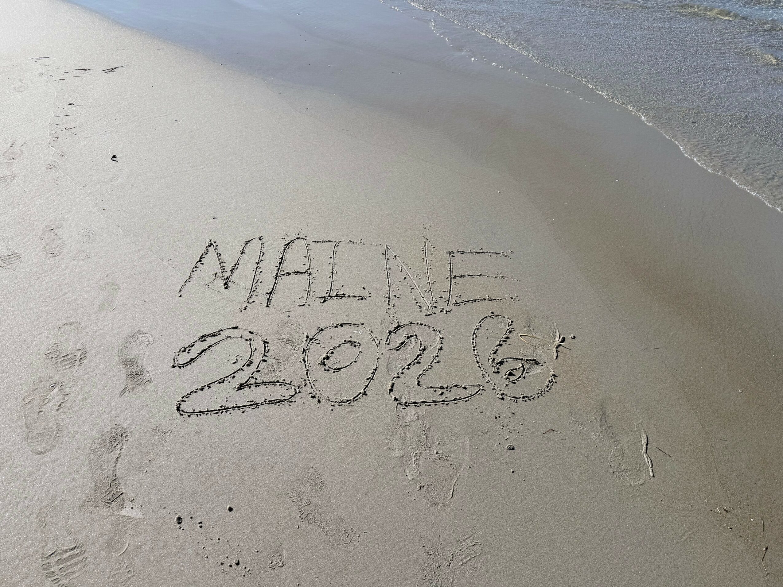 Words and numbers written in the sand on a beach, with water nearby, reading 'MALIE 2026'.