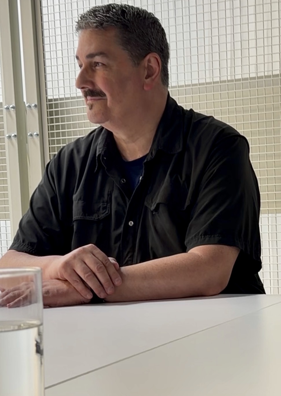 Man with dark hair and mustache sitting at a table, wearing a black shirt, with a glass of water nearby.