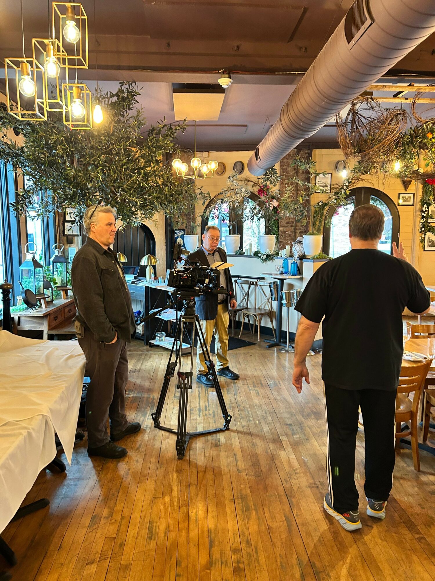 Two men talking indoors near a table, with a decorated indoor space and people in the background.