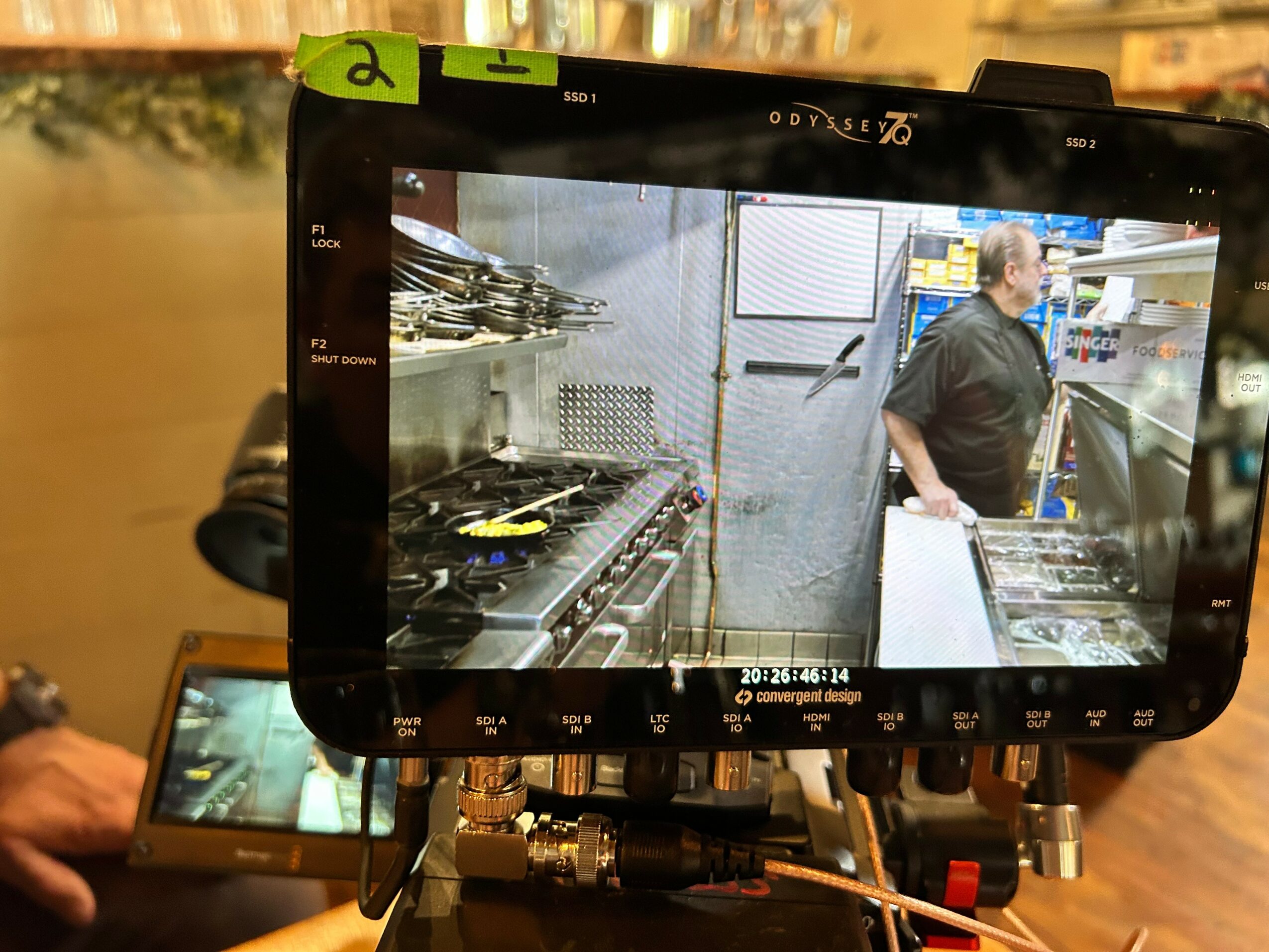 A camera monitor displays a person in a kitchen, with shelves and cooking equipment visible in the background.