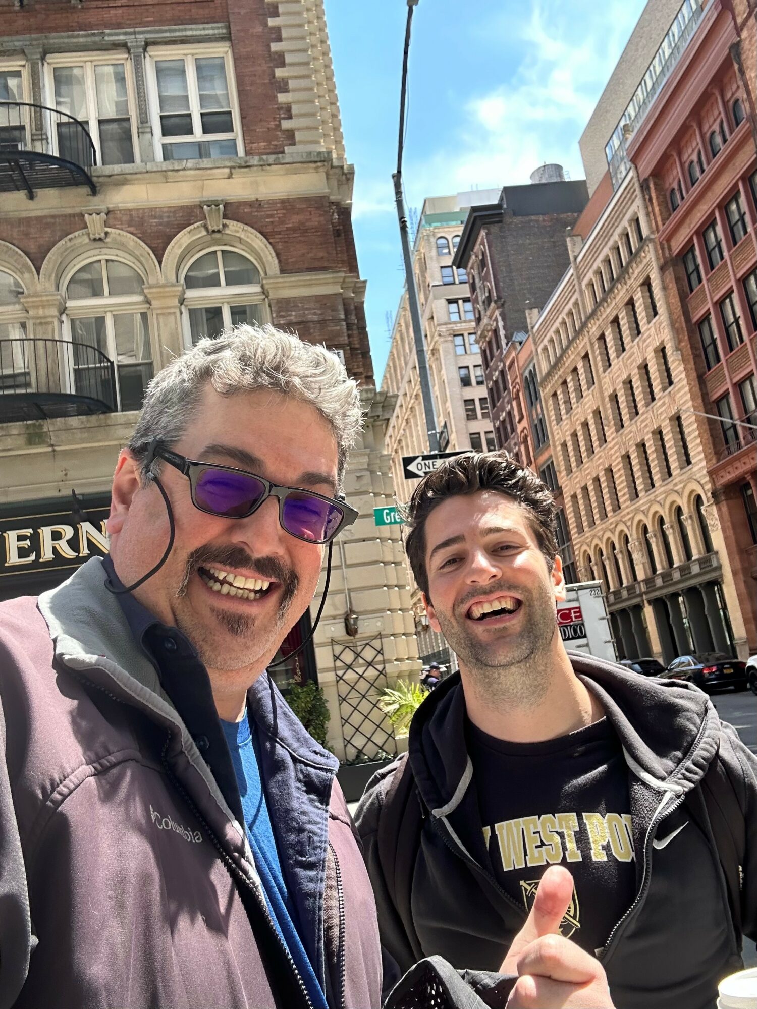 Two smiling men taking a selfie in an urban street with tall buildings and a blue sky.