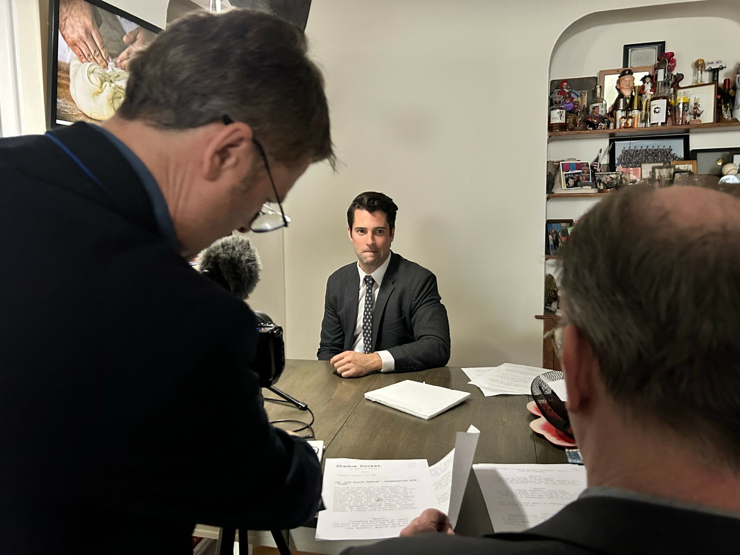 Man in suit sitting at table with papers, facing camera, in a room with shelves and a TV.