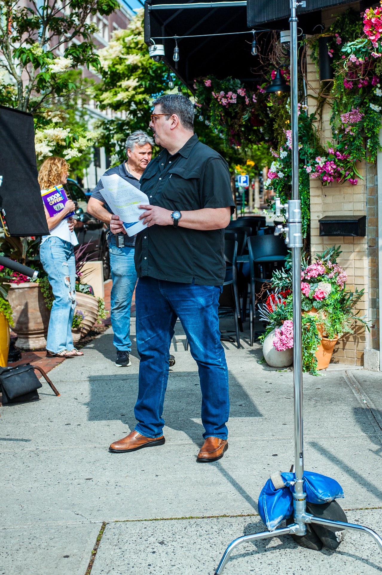 Man holding papers standing outdoors with people and flowers around, sunlight, and a blue bag on the ground.