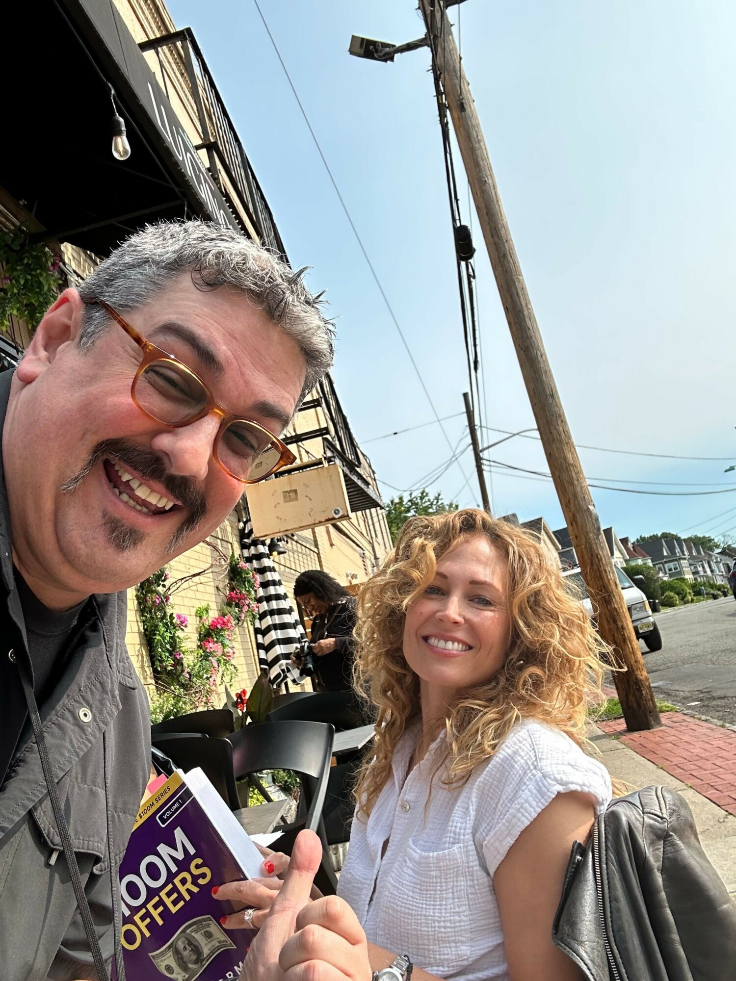 Two people smiling outdoors, one holding a microphone, with a building, power pole, and sky in the background.