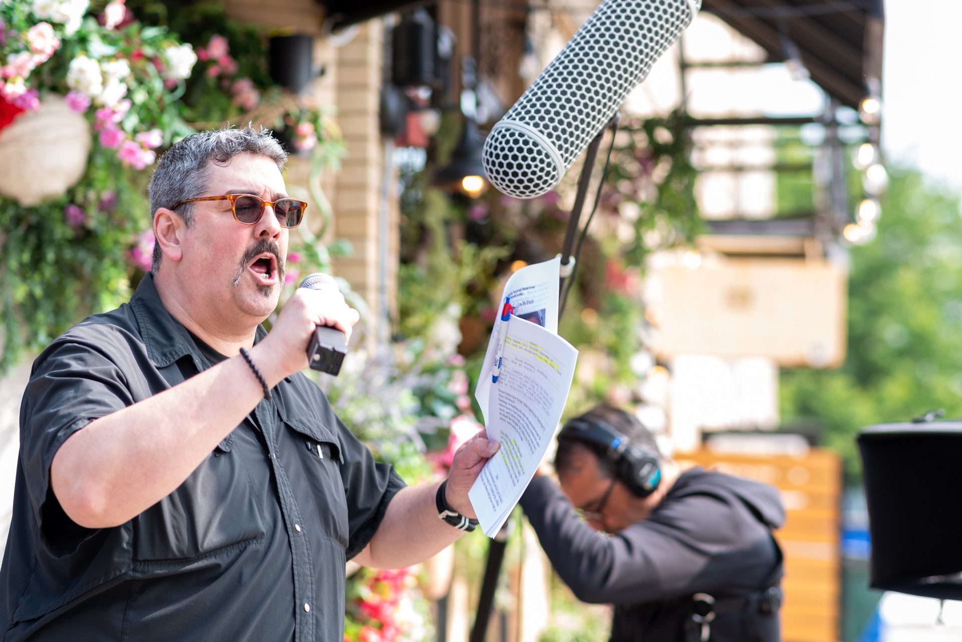 Man speaking into a microphone outdoors, holding papers, with a woman in the background wearing headphones, surrounded by flowers.