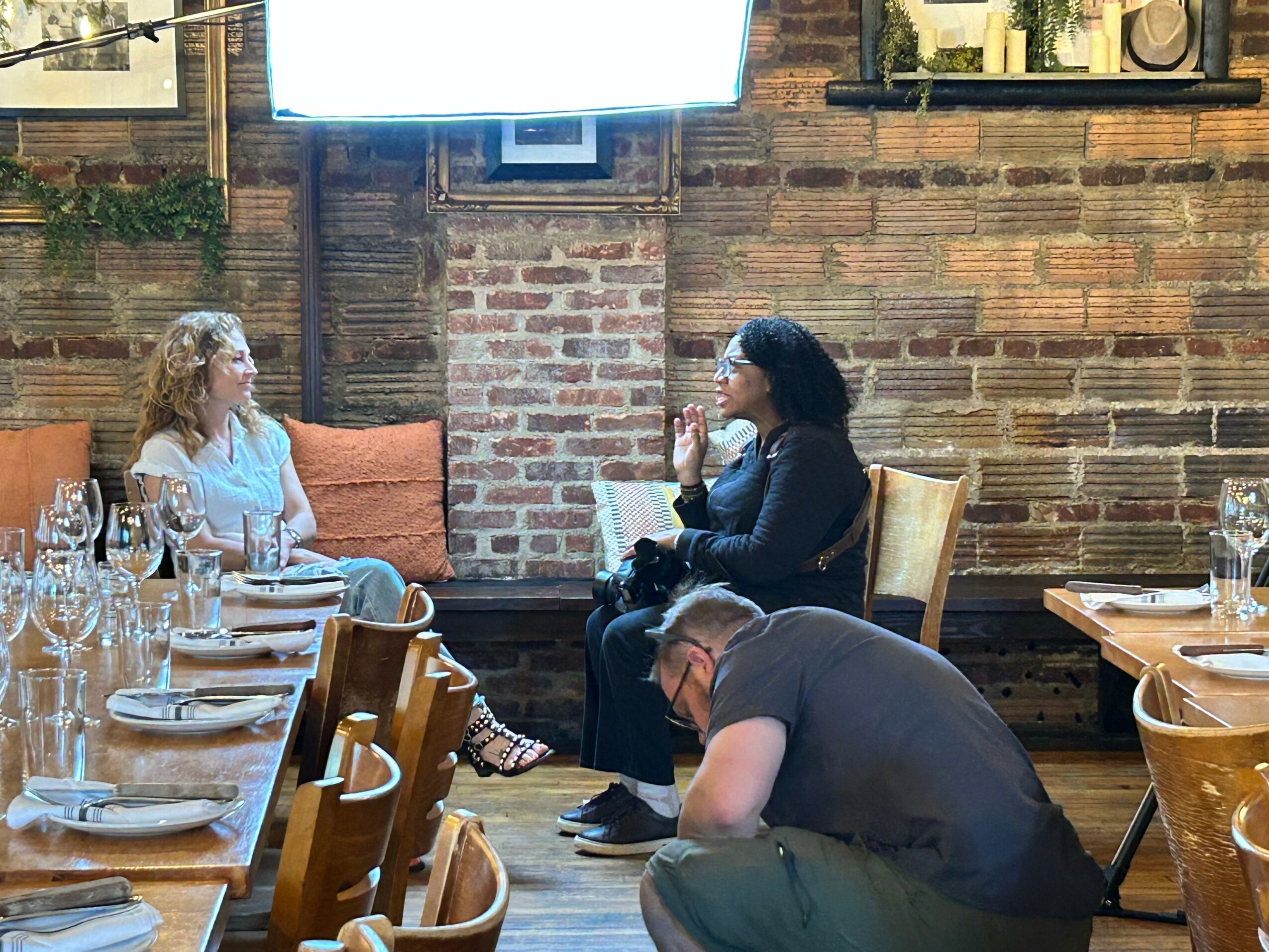 Two women sitting at a table in a restaurant, engaged in conversation, with a brick wall background and a large screen above.