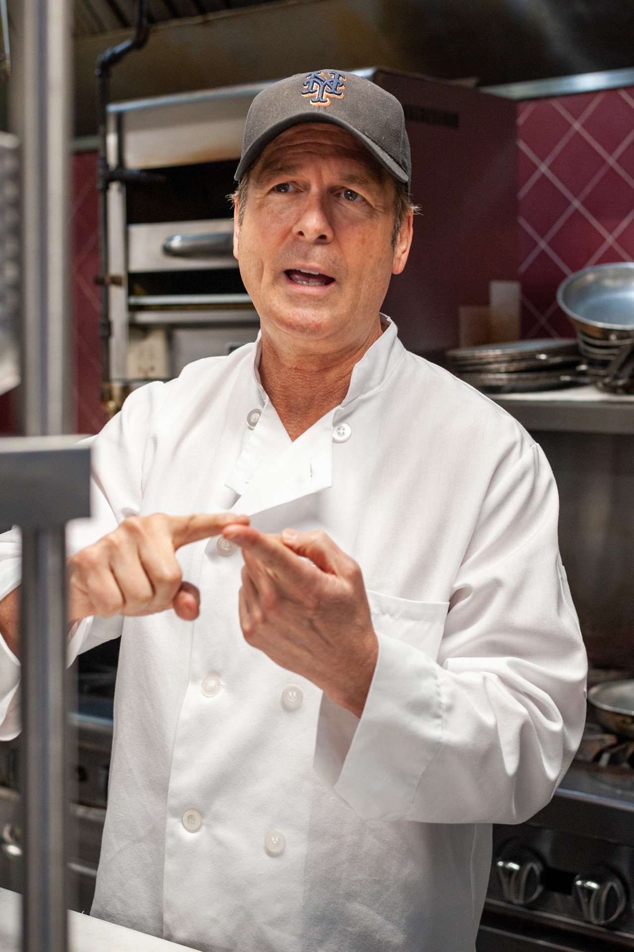 Chef in white uniform and cap speaking in a kitchen with shelves and dishes behind him.