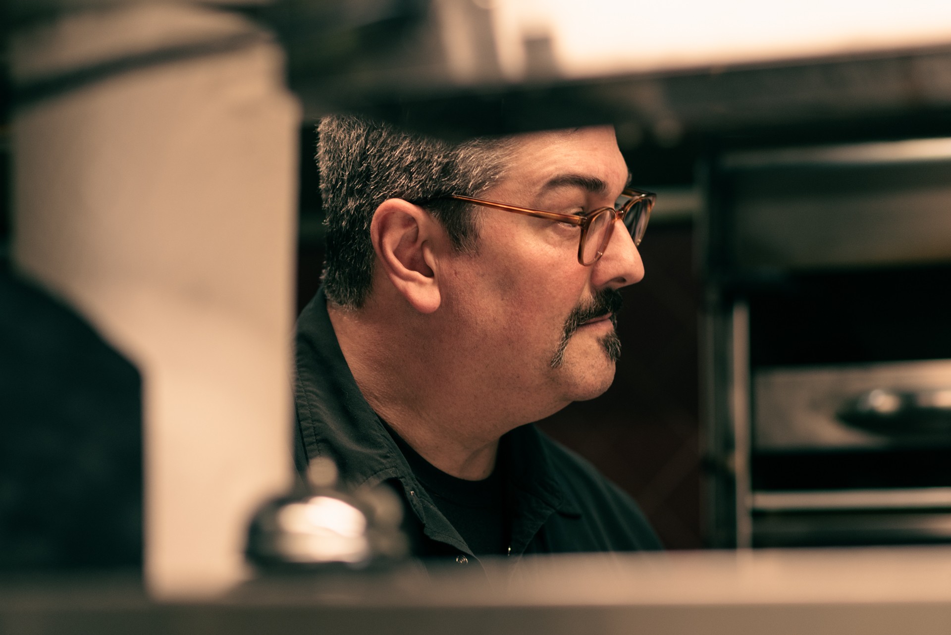Side profile of a man with glasses and a mustache, looking to the right, in an indoor setting.
