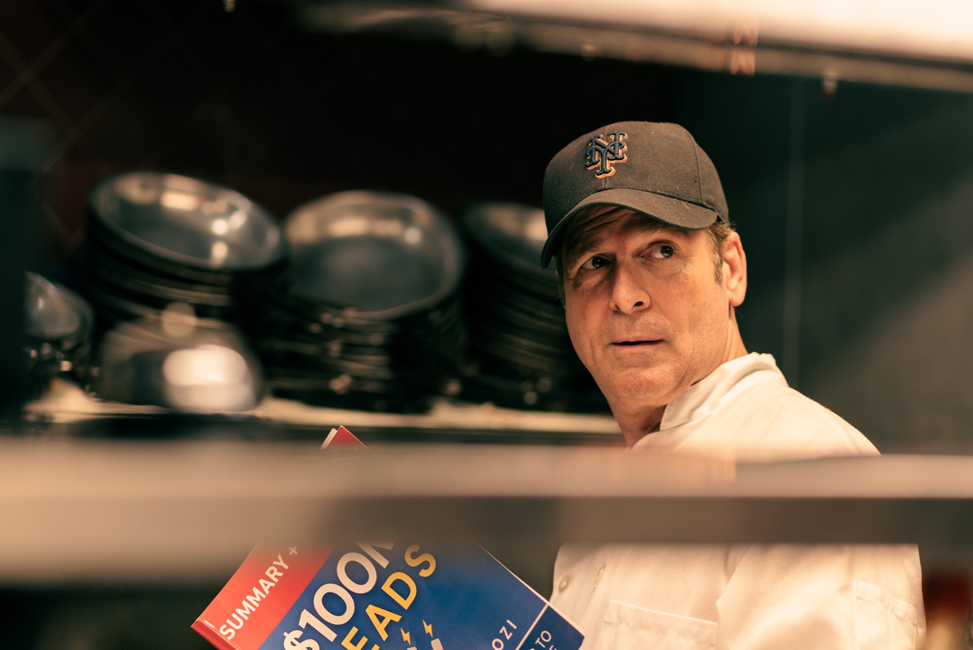 Man wearing a cap looking to the side behind a shelf of plates, with a sign reading 'SUMMAR 100 LEADS'.