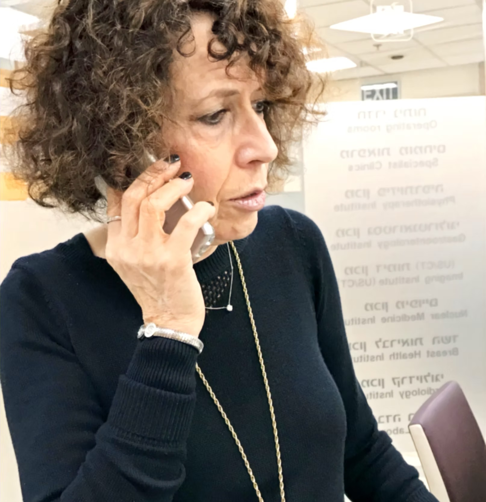 Woman with curly hair talking on a phone indoors, wearing a dark sweater and a long necklace.