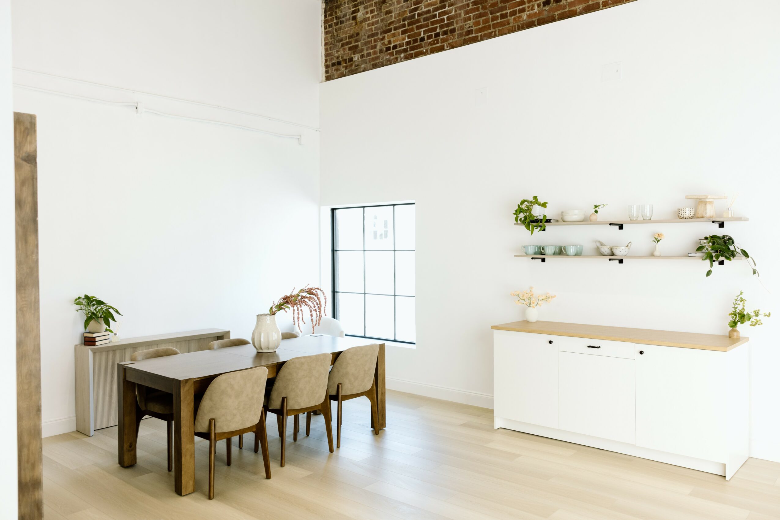 Bright dining room with white walls, a window, a dark wooden table, four beige chairs, and white shelves with plants.