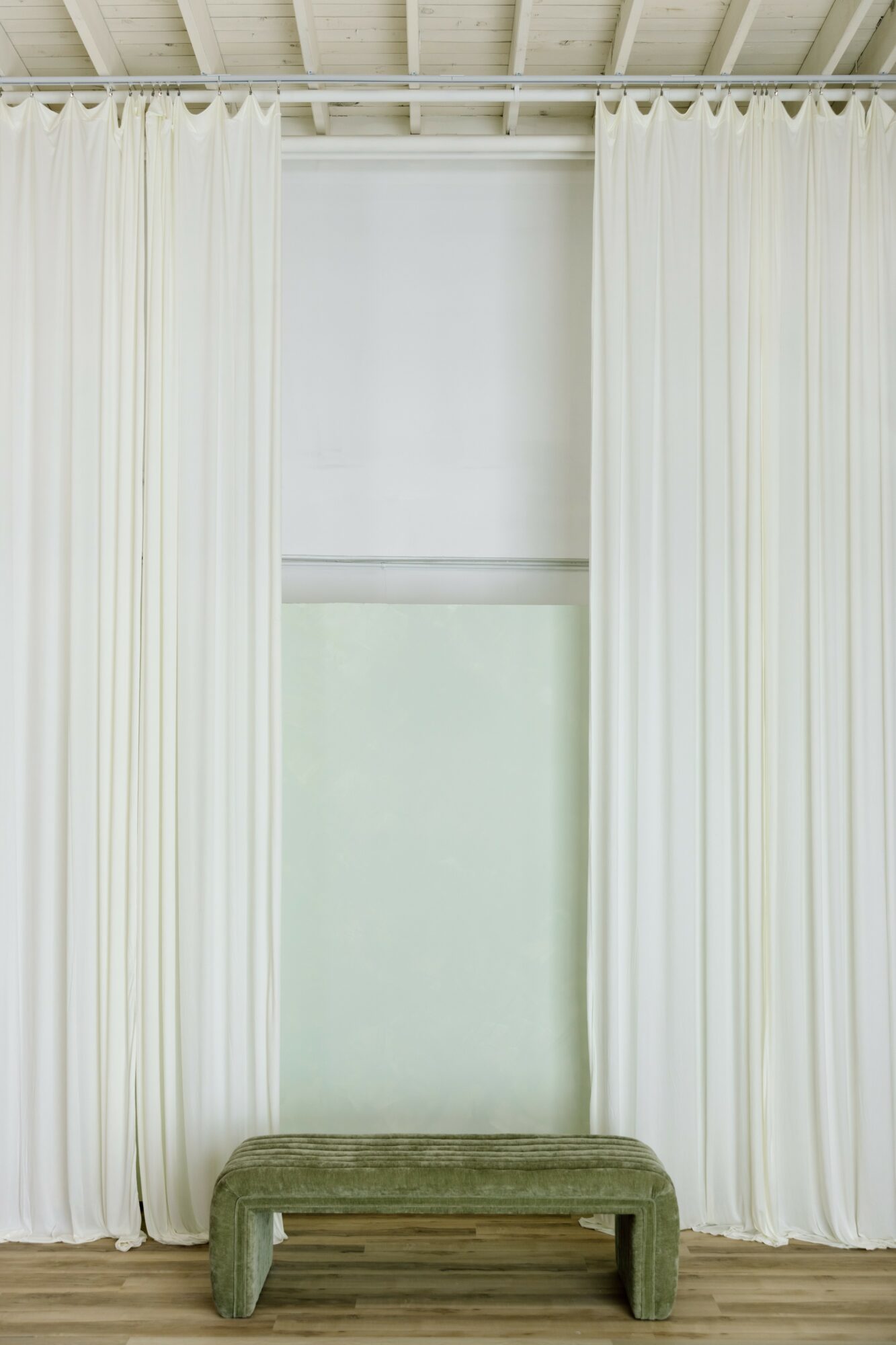 Window with white curtains and a green bench in front, wooden floor, and a white wall behind the curtains.