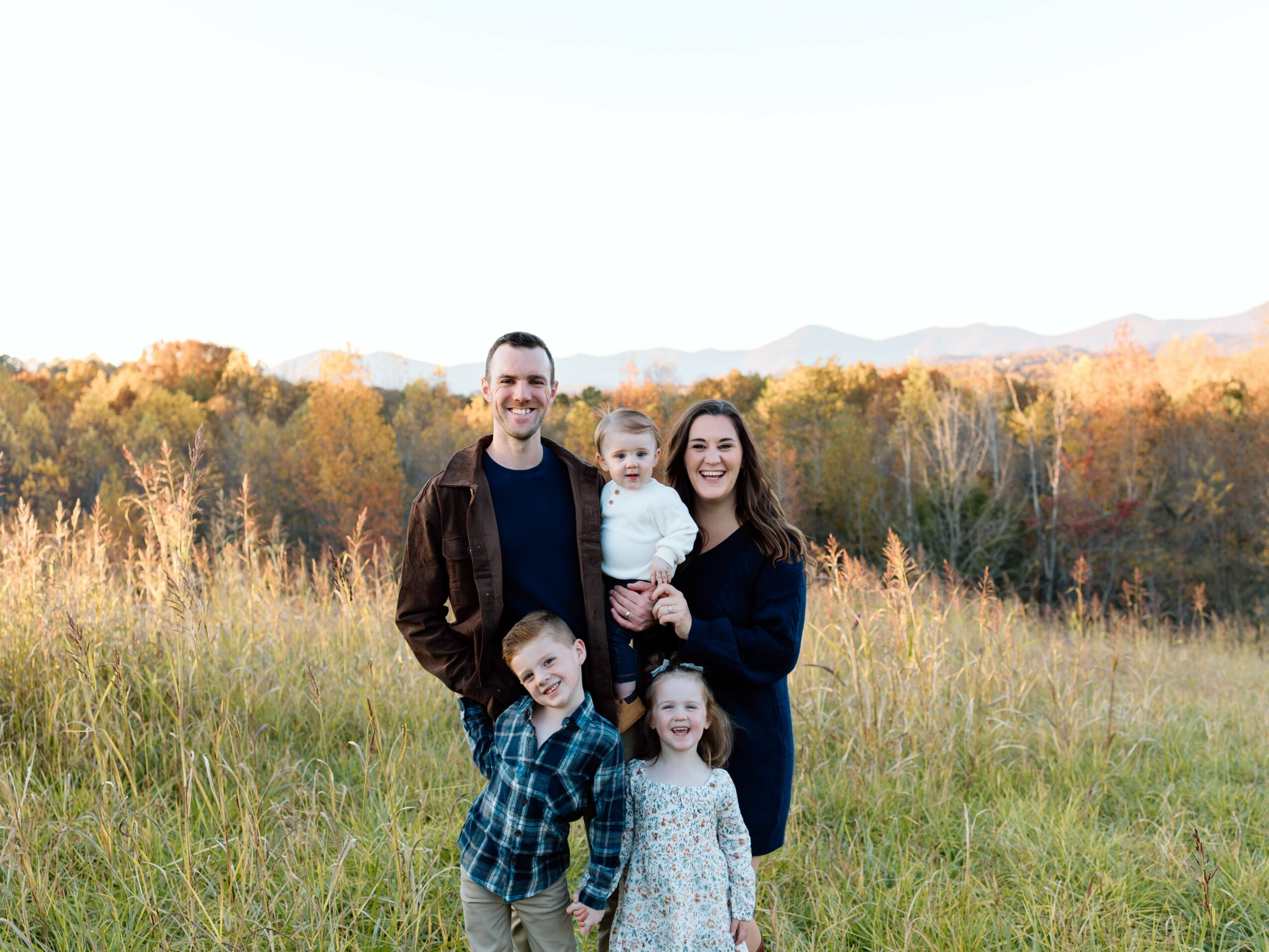 Family of four standing in a field with trees and mountains in the background, smiling outdoors during daytime.