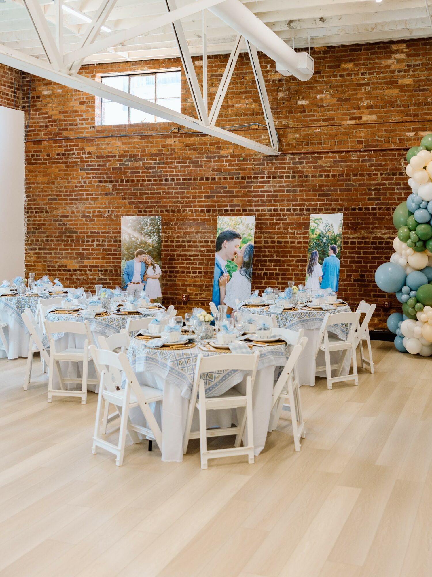 Event space with round tables, white chairs, brick wall, and balloon decoration, set for a celebration or gathering.