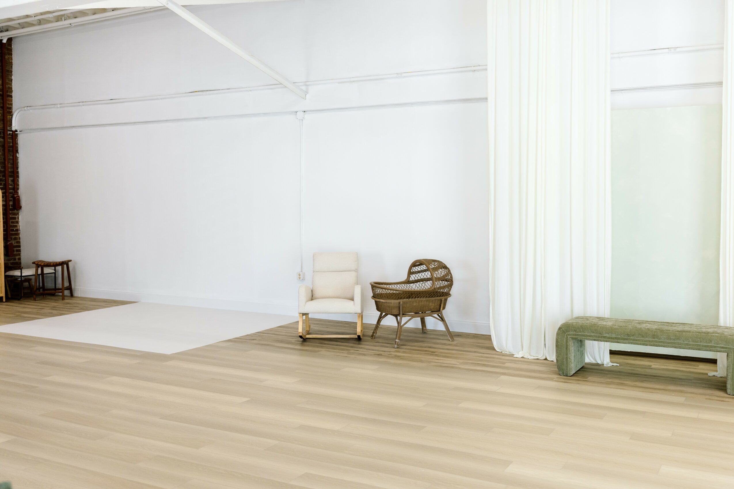 Empty room with white walls, wooden floor, and minimal furniture including chairs, a bench, and curtains.
