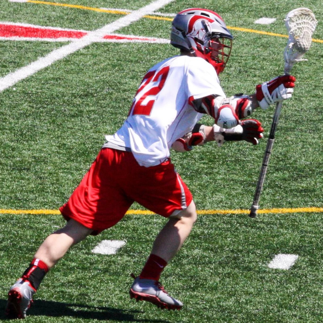 Young lacrosse player in white and red uniform running on field with stick and helmet, holding lacrosse ball, grass background.