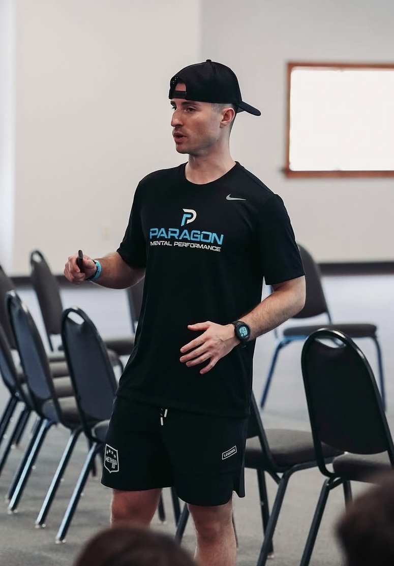 Young man in athletic clothing and cap standing in a room with chairs, holding a pen, with a whiteboard behind him.