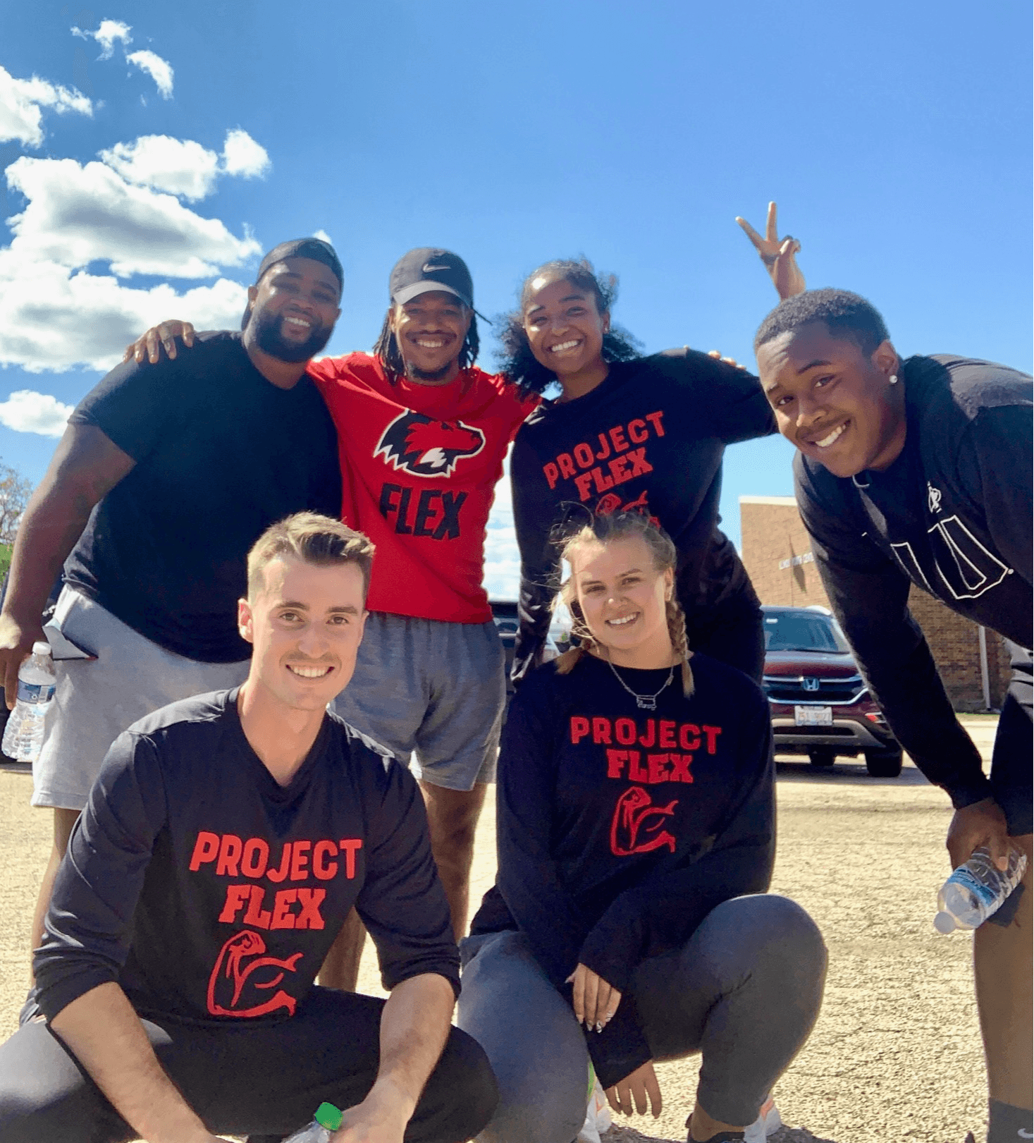 Group of six smiling people outdoors, some making peace signs, under a blue sky with clouds.