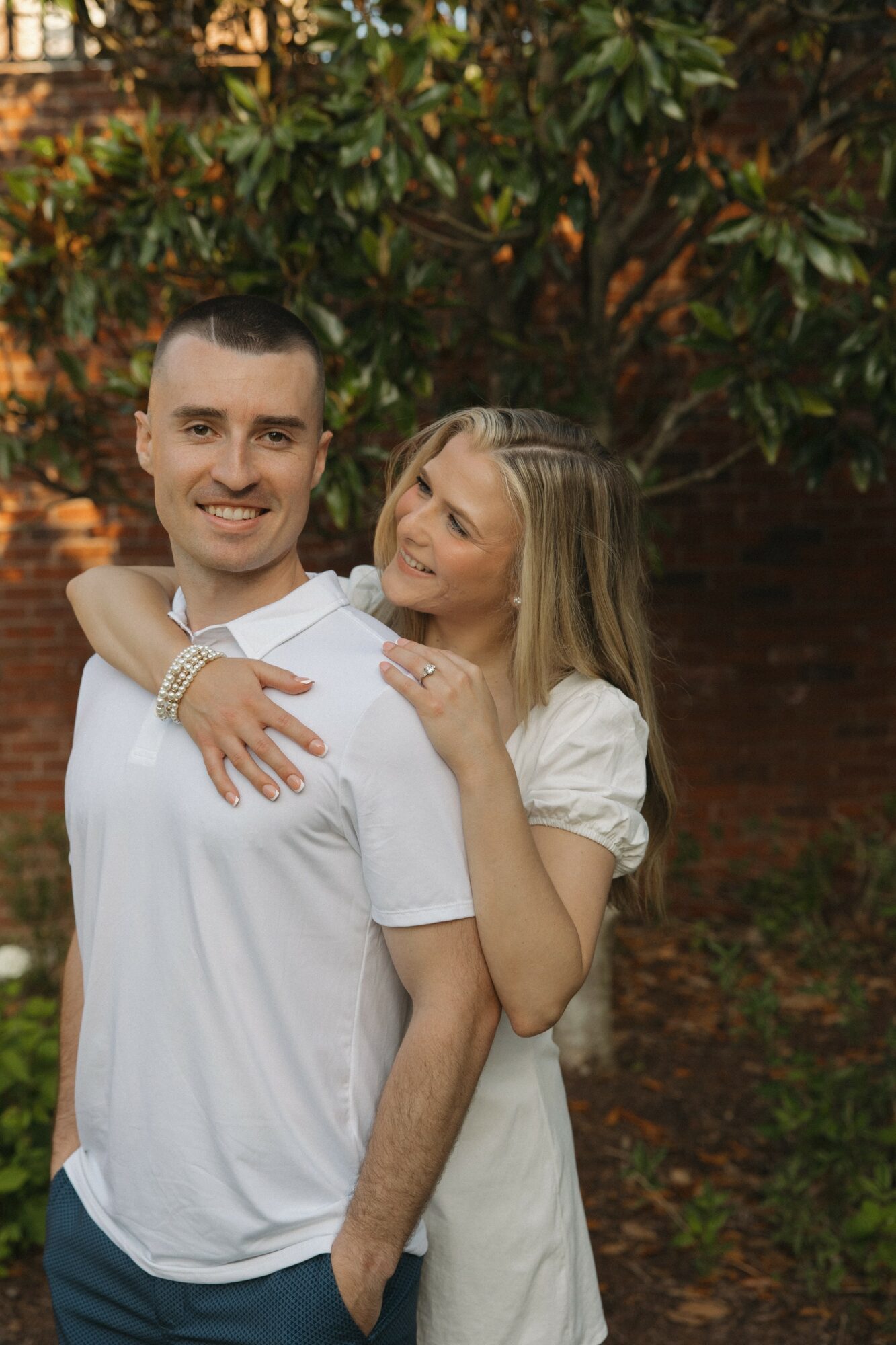 A smiling man and woman outdoors, woman hugging man from behind, trees and brick wall in background.
