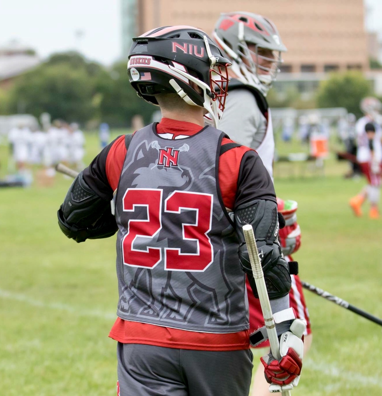 Lacrosse player wearing a helmet and jersey with number 23, holding a lacrosse stick on a grassy field.