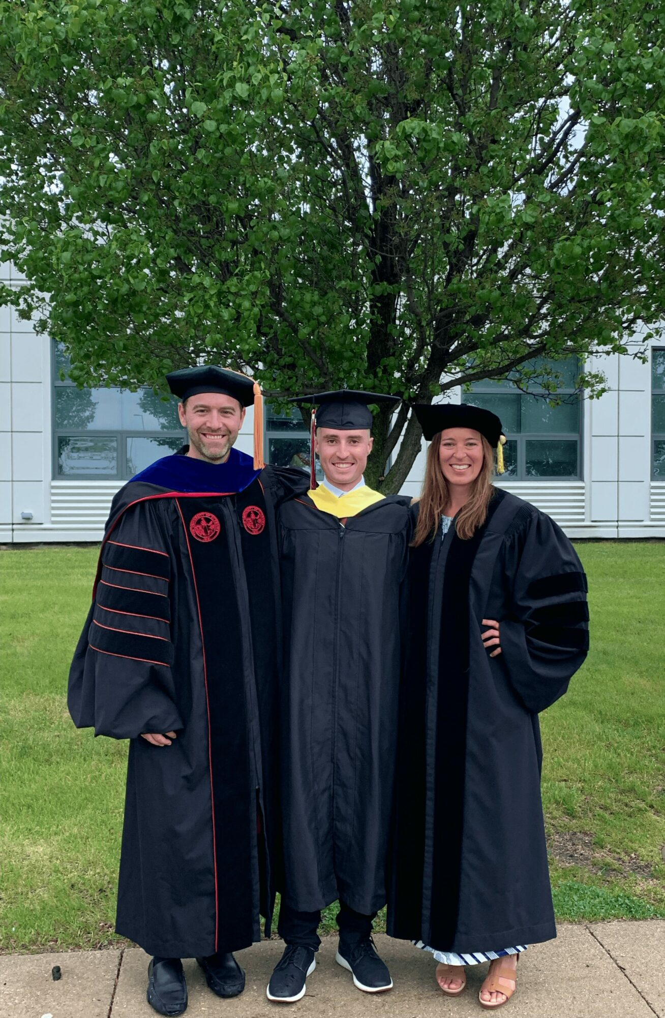 Three graduates in caps and gowns standing outdoors in front of a tree and building, smiling for the photo.