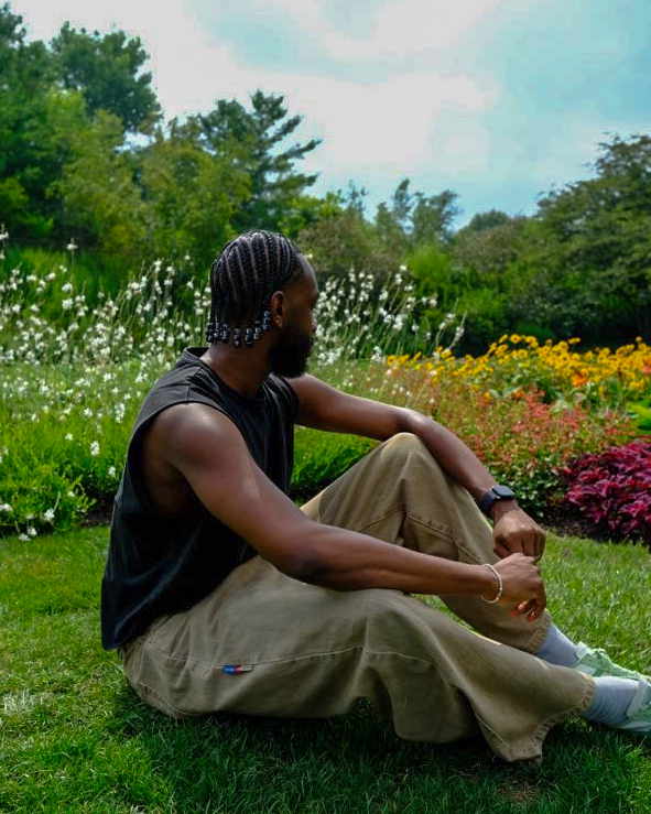 Person sitting on grass in a garden with colorful flowers and trees, facing left, with arms resting on knees.