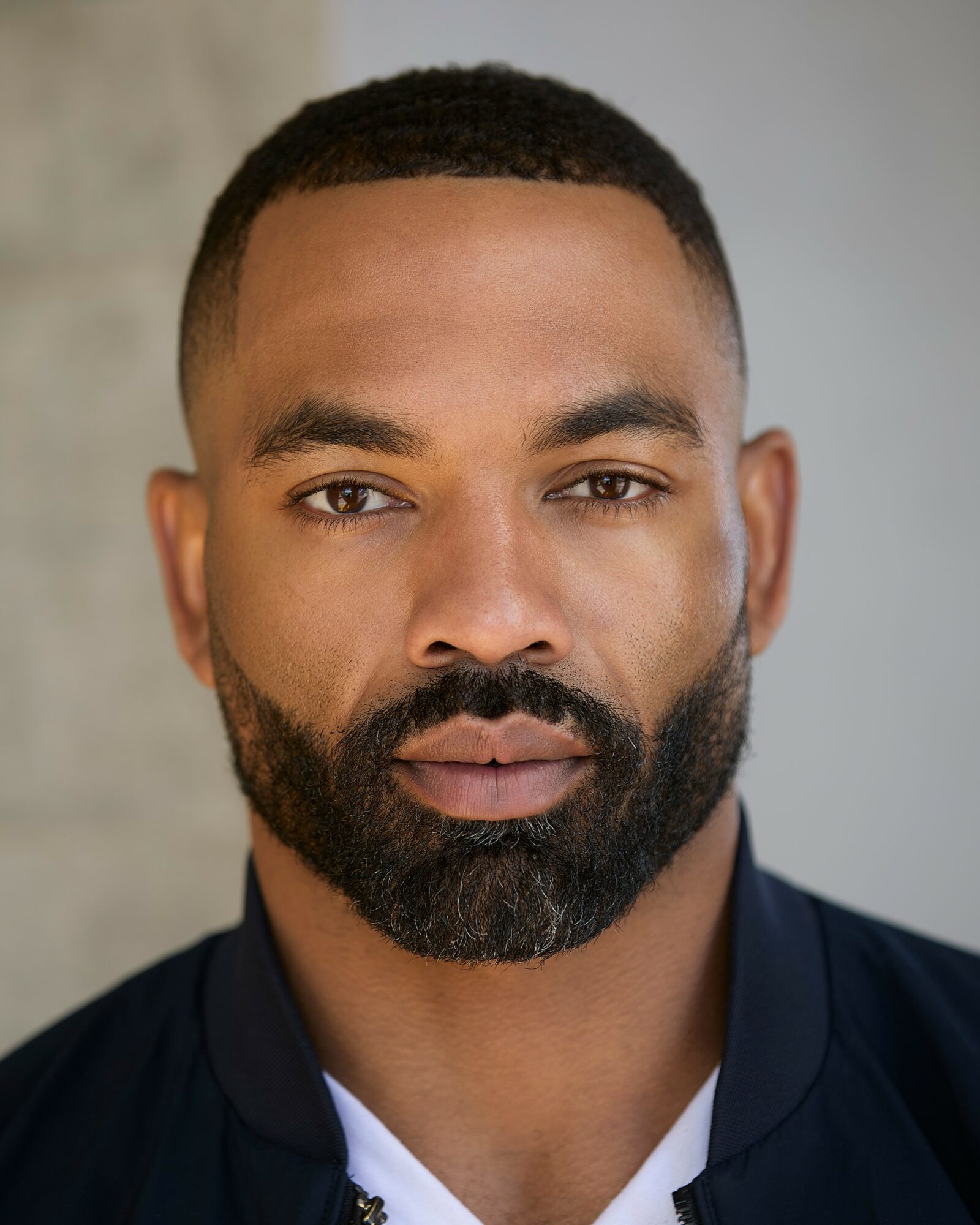 Close-up portrait of a man with short hair and a beard, wearing a dark jacket and white shirt, neutral background.